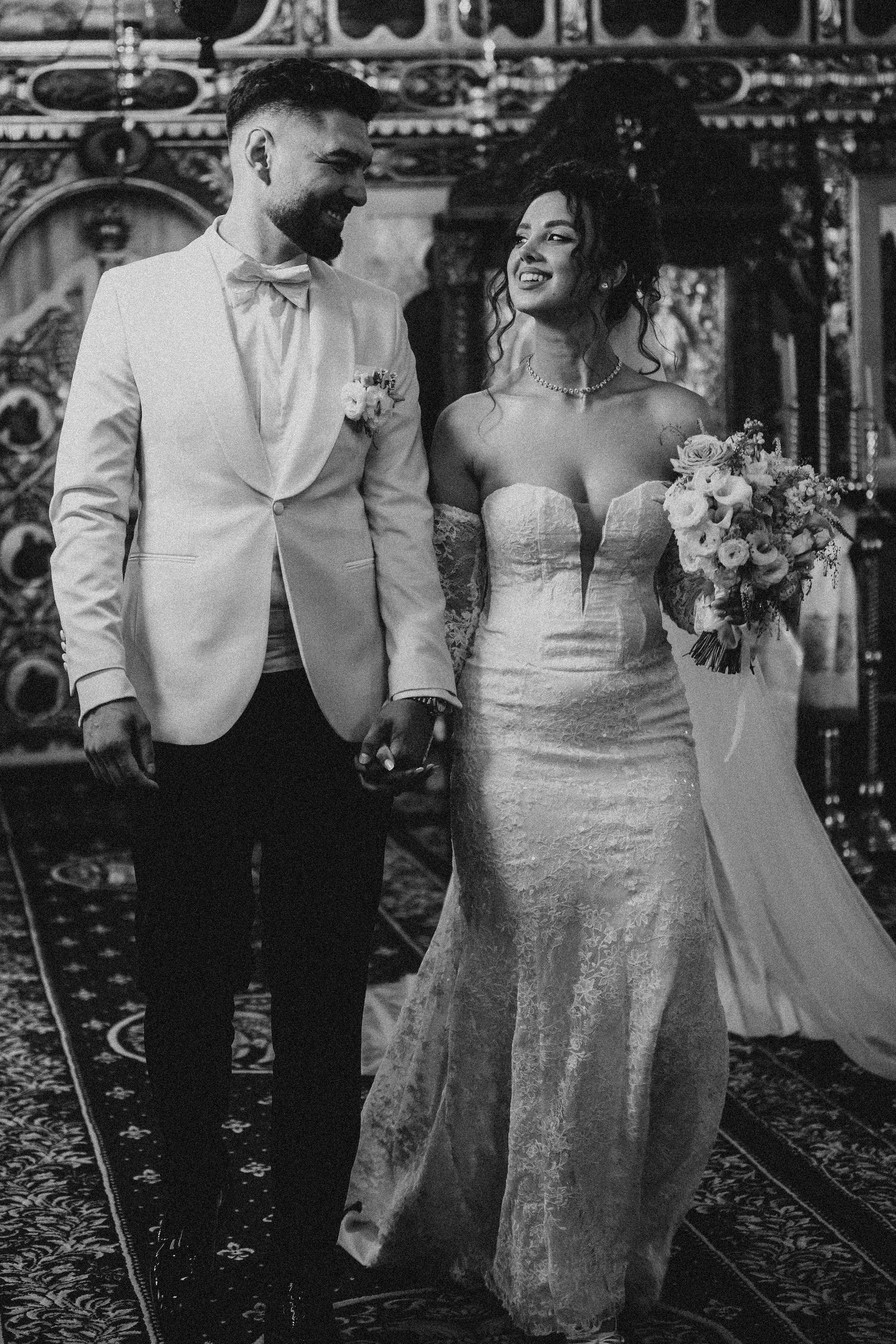 Black and white photo of a smiling bride in strapless lace gown holding bouquet walking with groom in white tuxedo.