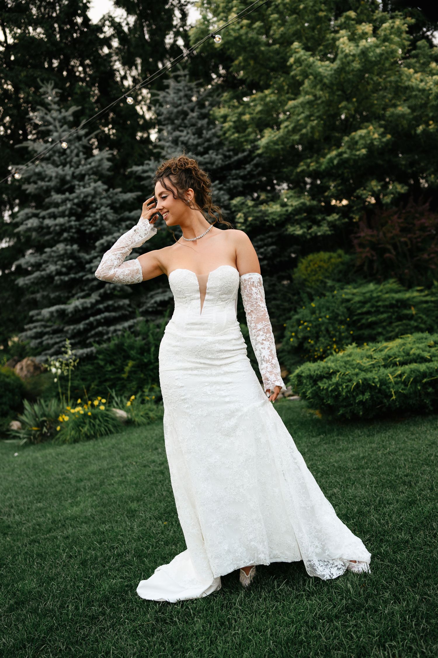 Bride in a strapless lace wedding gown with long lace gloves standing on green grass with trees in background.