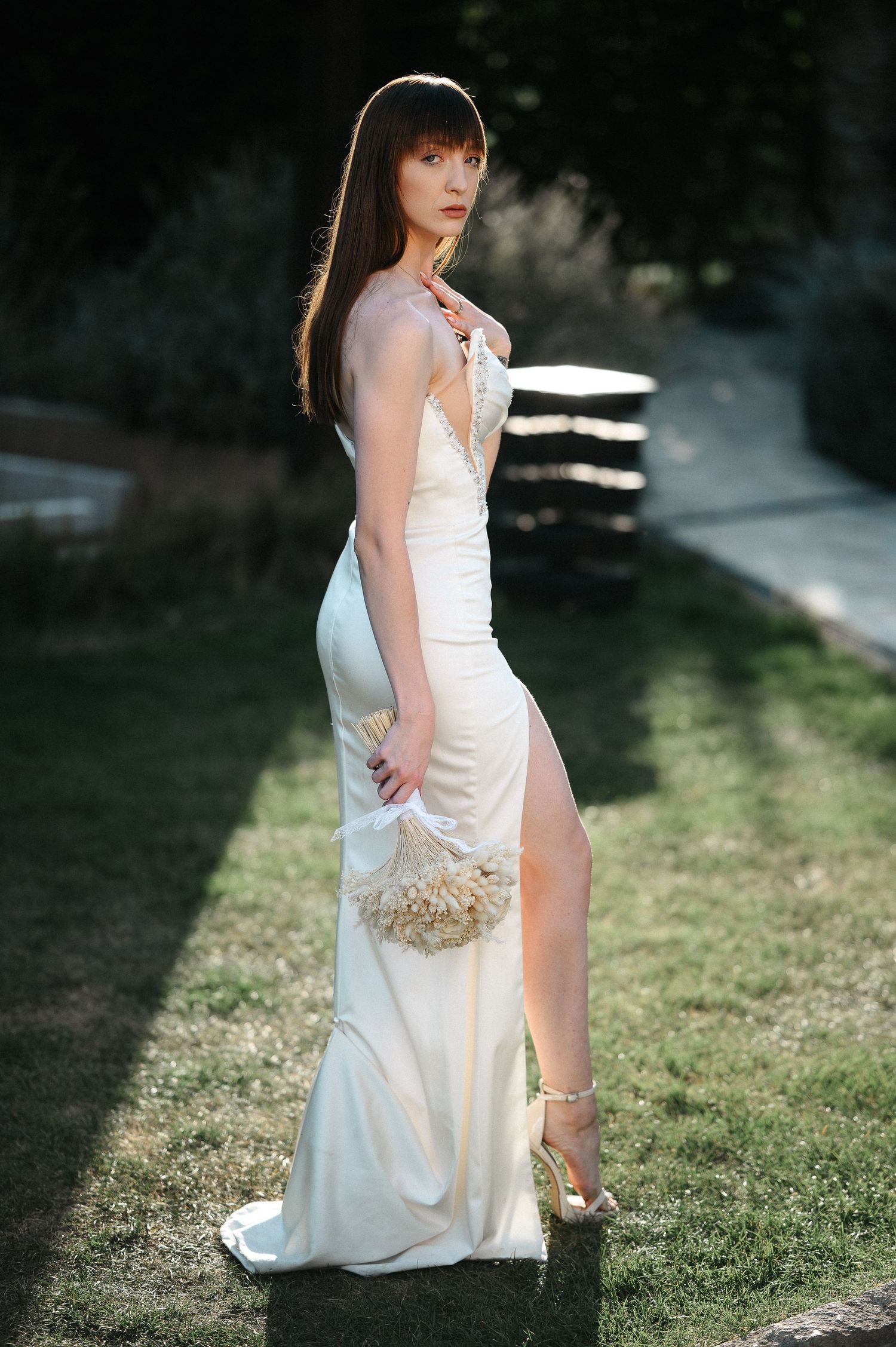 Bride in a fitted white slit dress holding a floral bouquet while standing on grass in a garden.