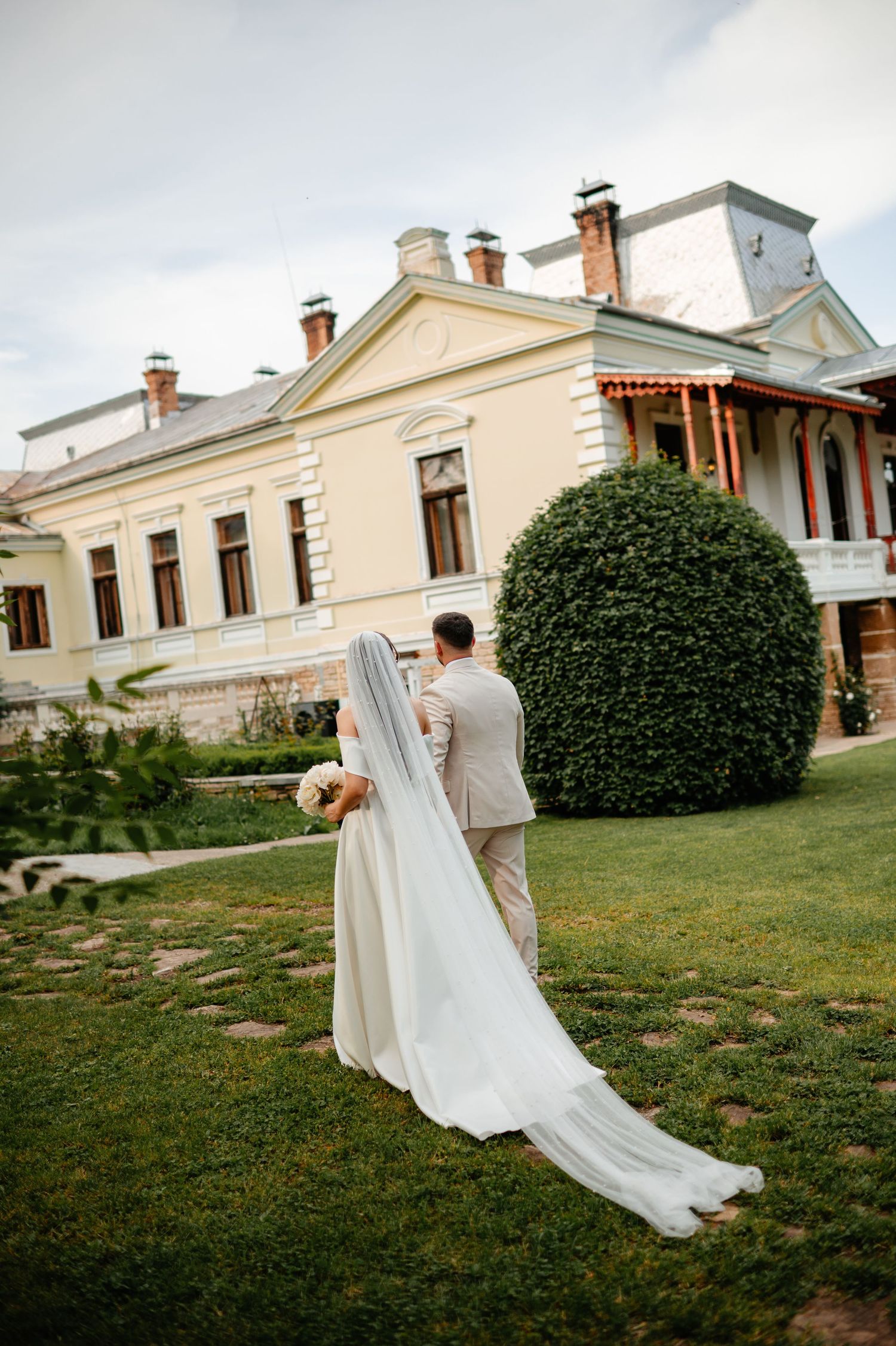 Bride and groom walk hand in hand toward elegant yellow manor house surrounded by lush green gardens on wedding day.