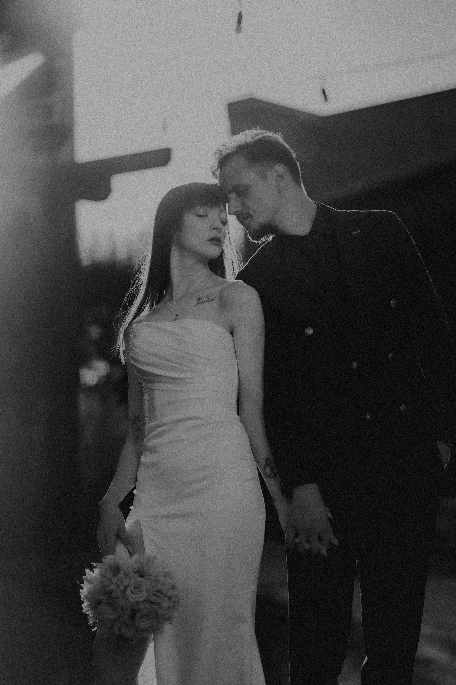 Black and white photo of bride and groom in intimate pose, she holds bouquet in elegant white dress.