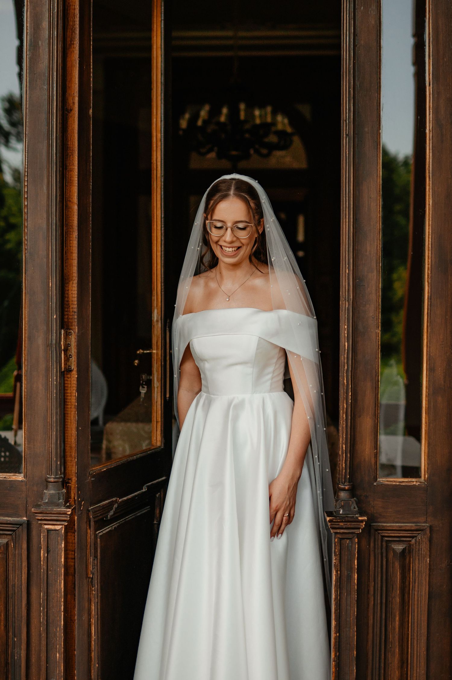 Smiling bride in off-shoulder white dress and veil stands in ornate wooden doorway outdoors.