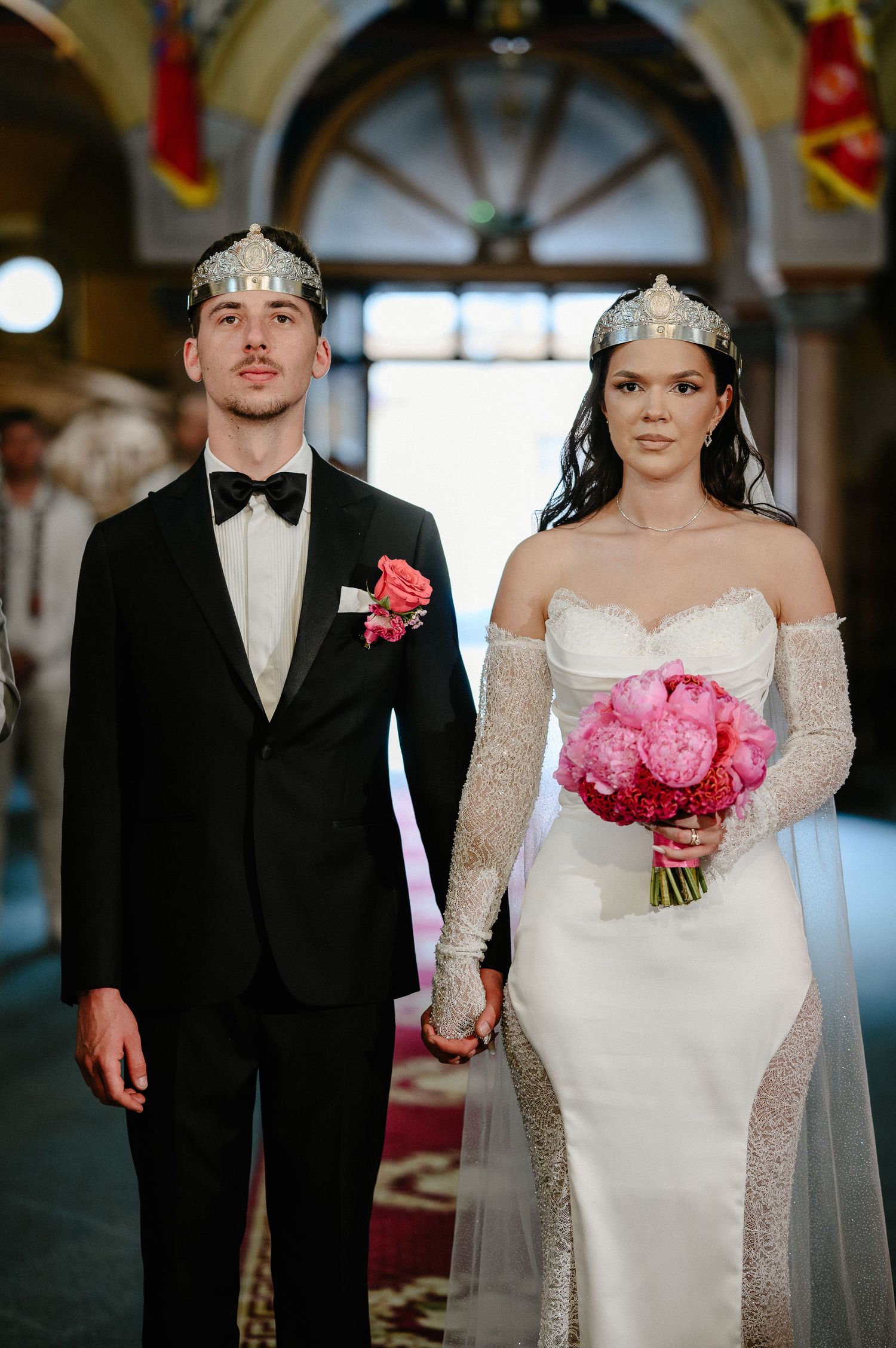 Bride and groom wear Orthodox wedding crowns, holding hands and pink bouquet in church aisle.