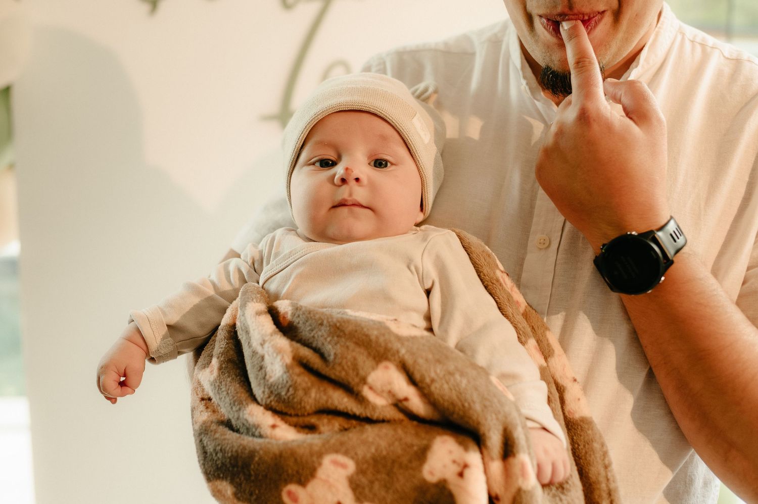 Adorable newborn baby in white outfit and bear hat being cradled by father indoors.