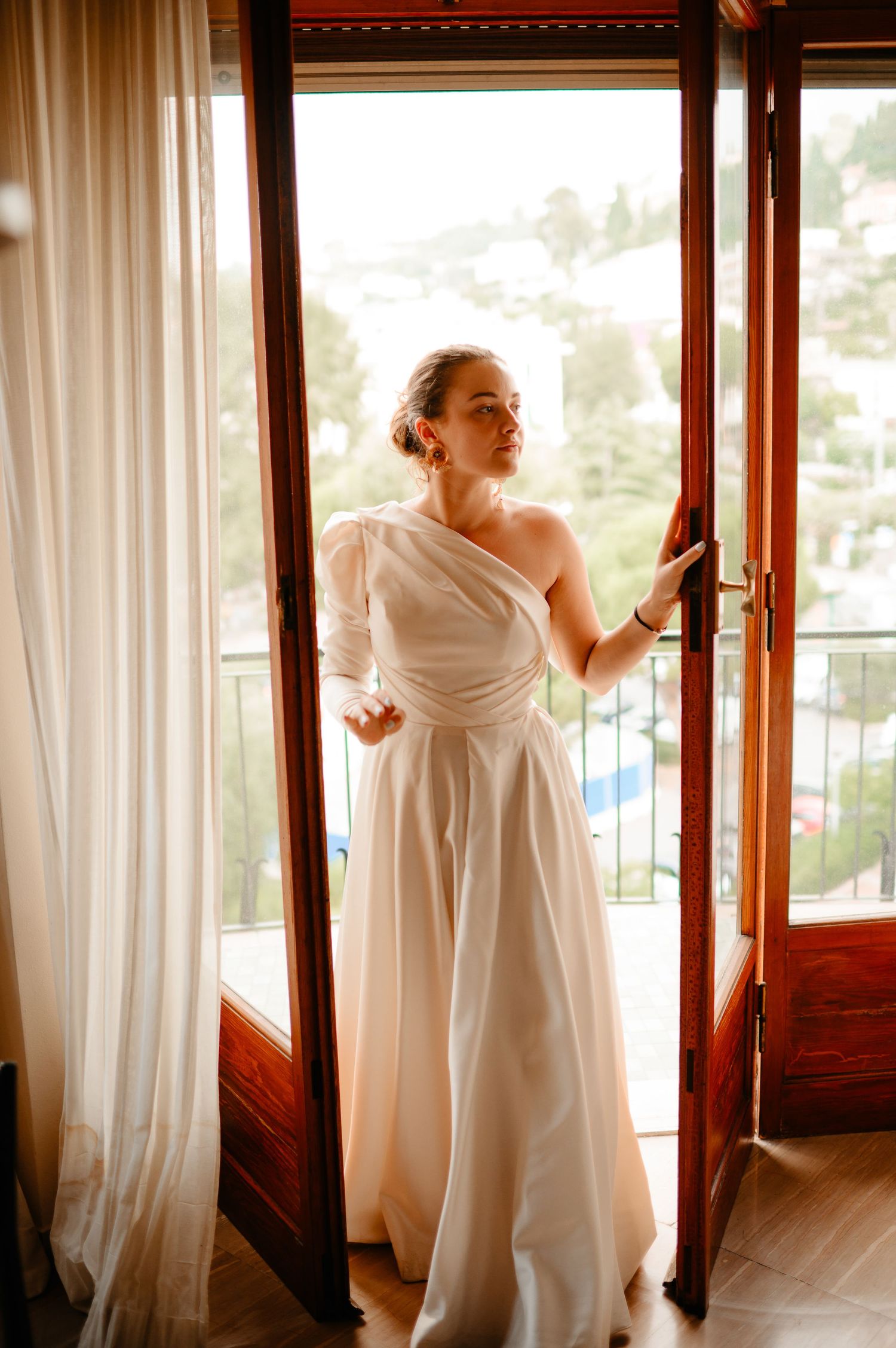 Elegant bride in asymmetric white gown standing in open French doors with garden view.