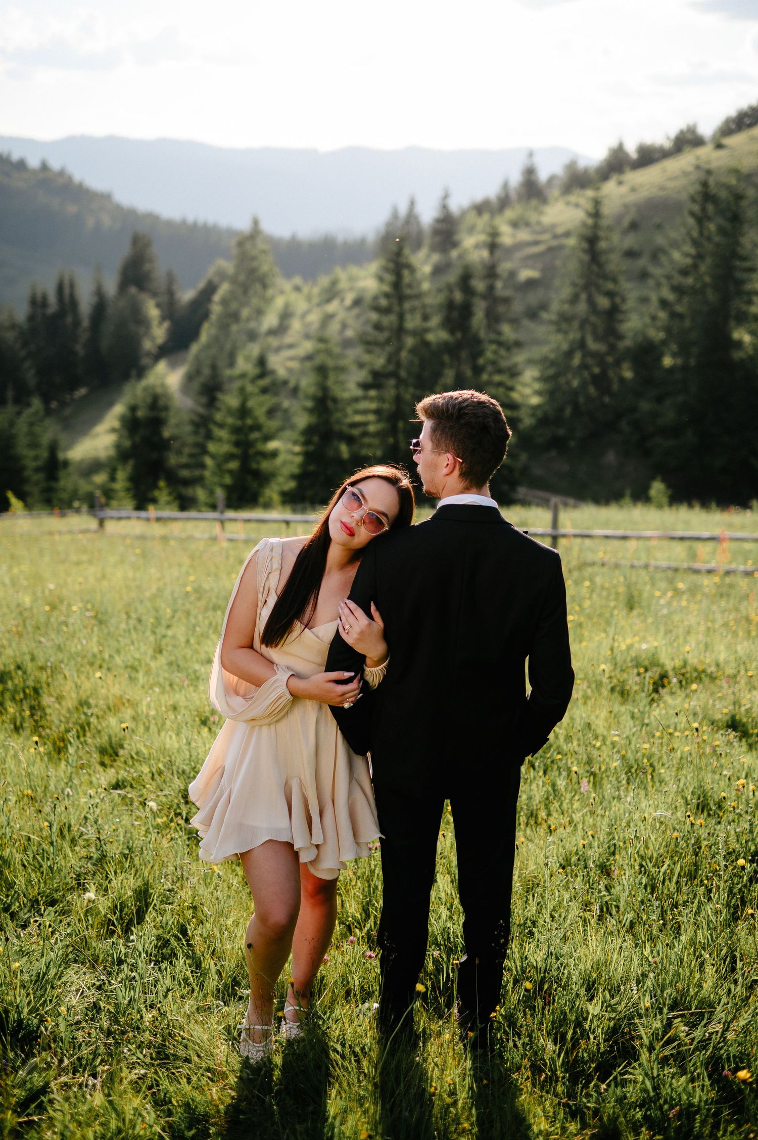 Couple embracing in a green mountain meadow, woman in cream dress leans on man in black suit.