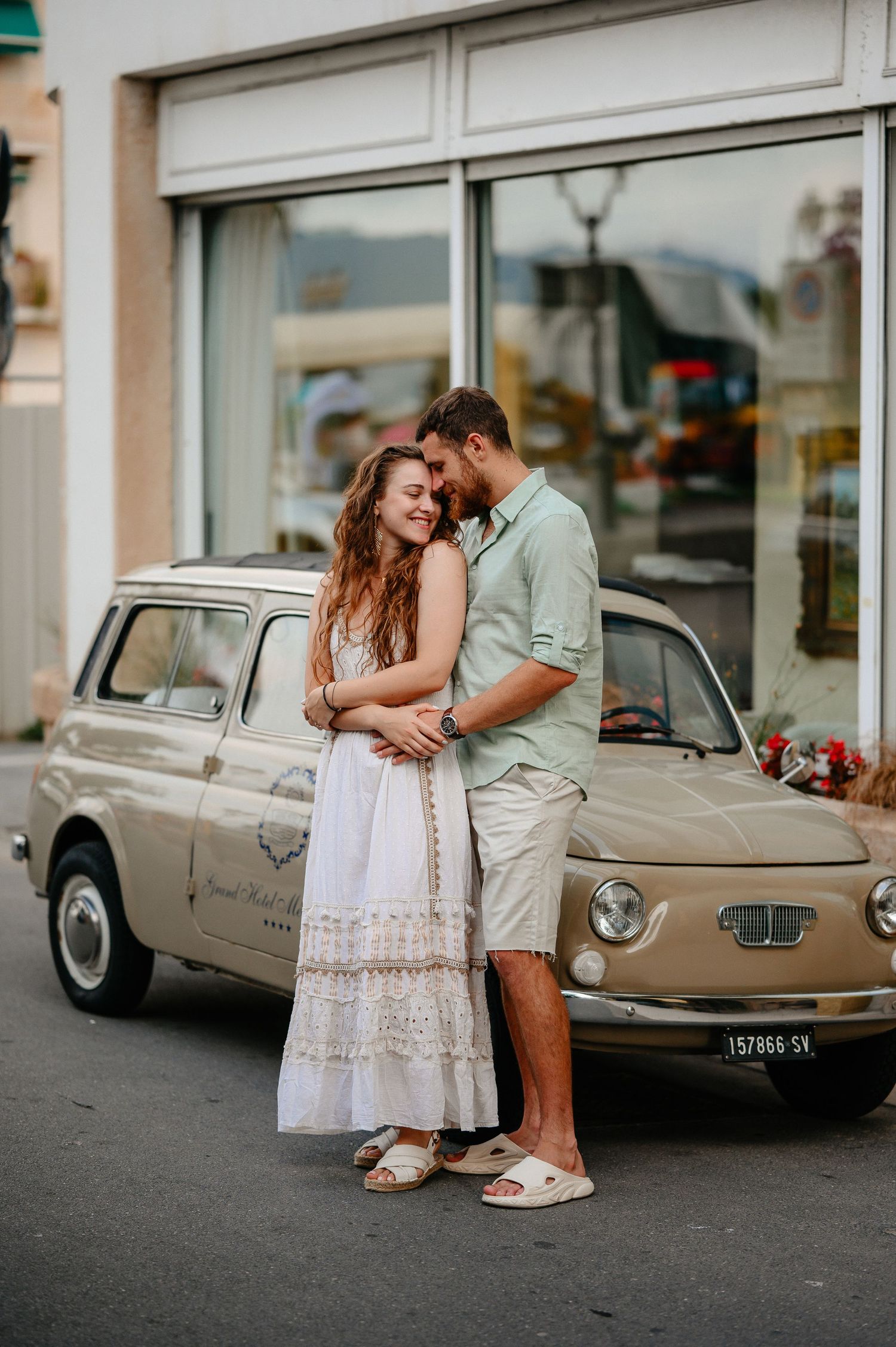 Romantic couple embracing in front of a vintage beige Fiat 500 on a charming street.