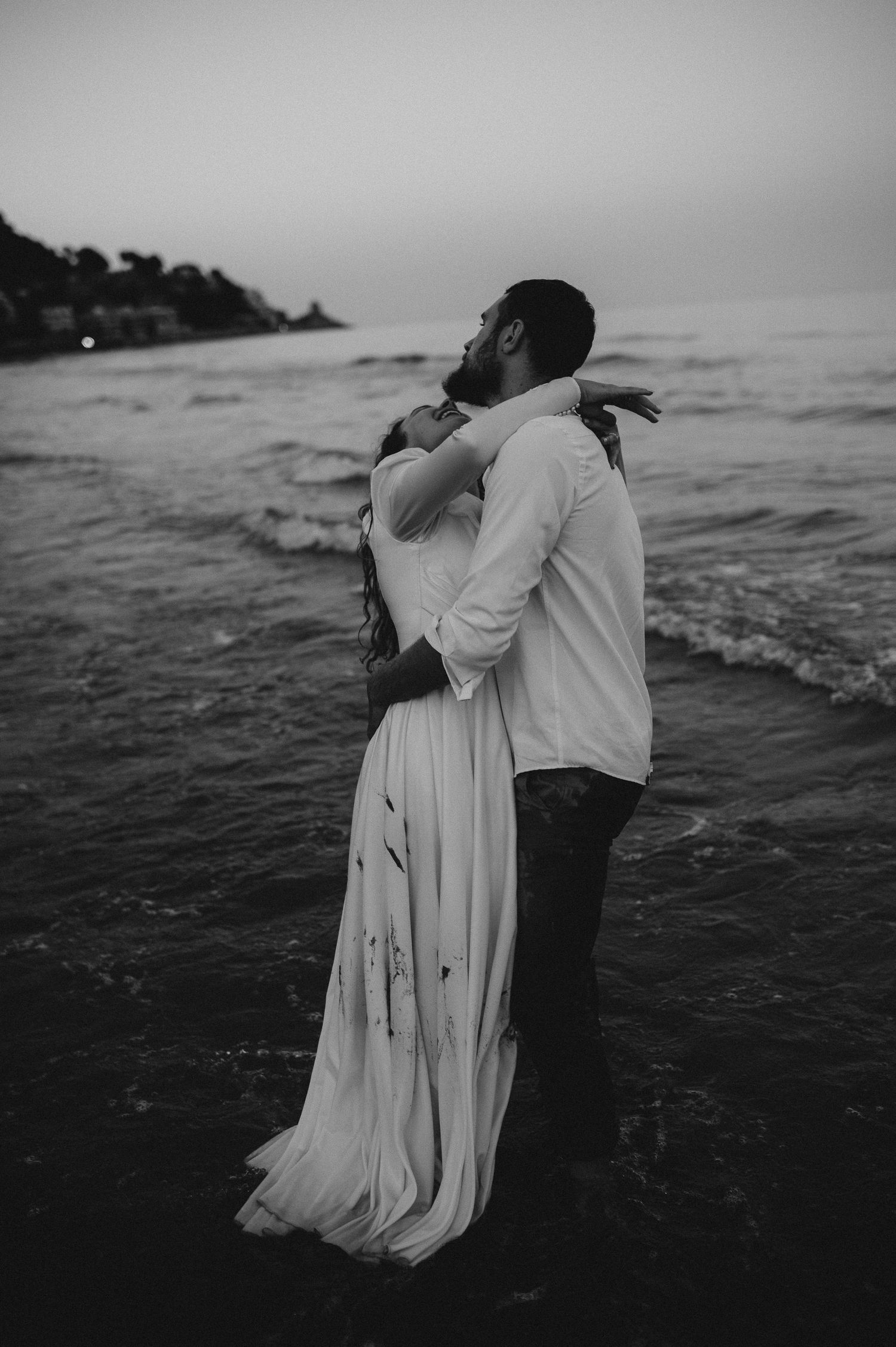 Black and white photo of couple embracing romantically in ocean waves at dusk on a sandy beach.