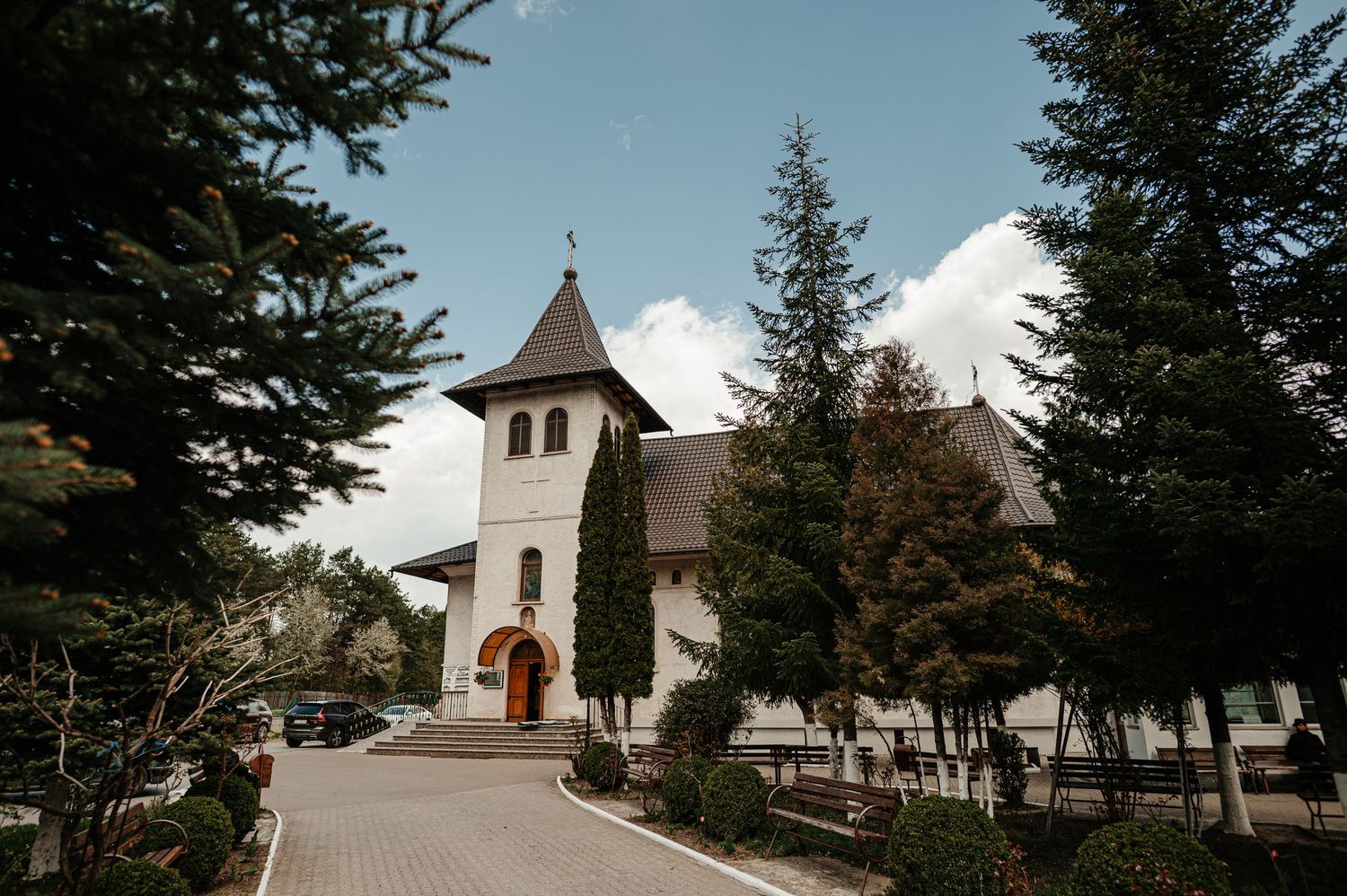 A white Orthodox church with a dark roof surrounded by tall pine trees under a cloudy sky.