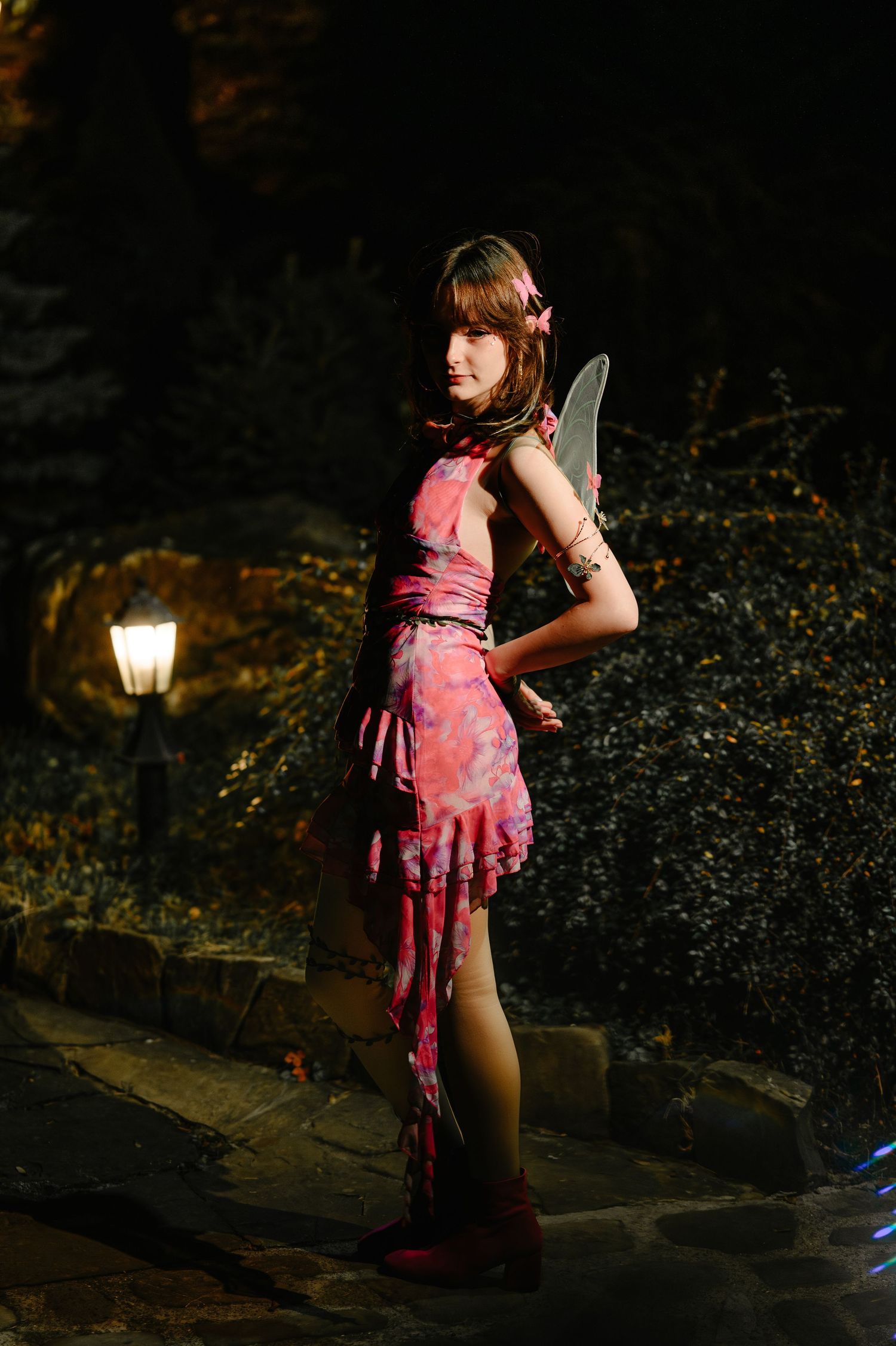 Girl in pink fairy costume with wings poses near glowing lantern in nighttime garden.