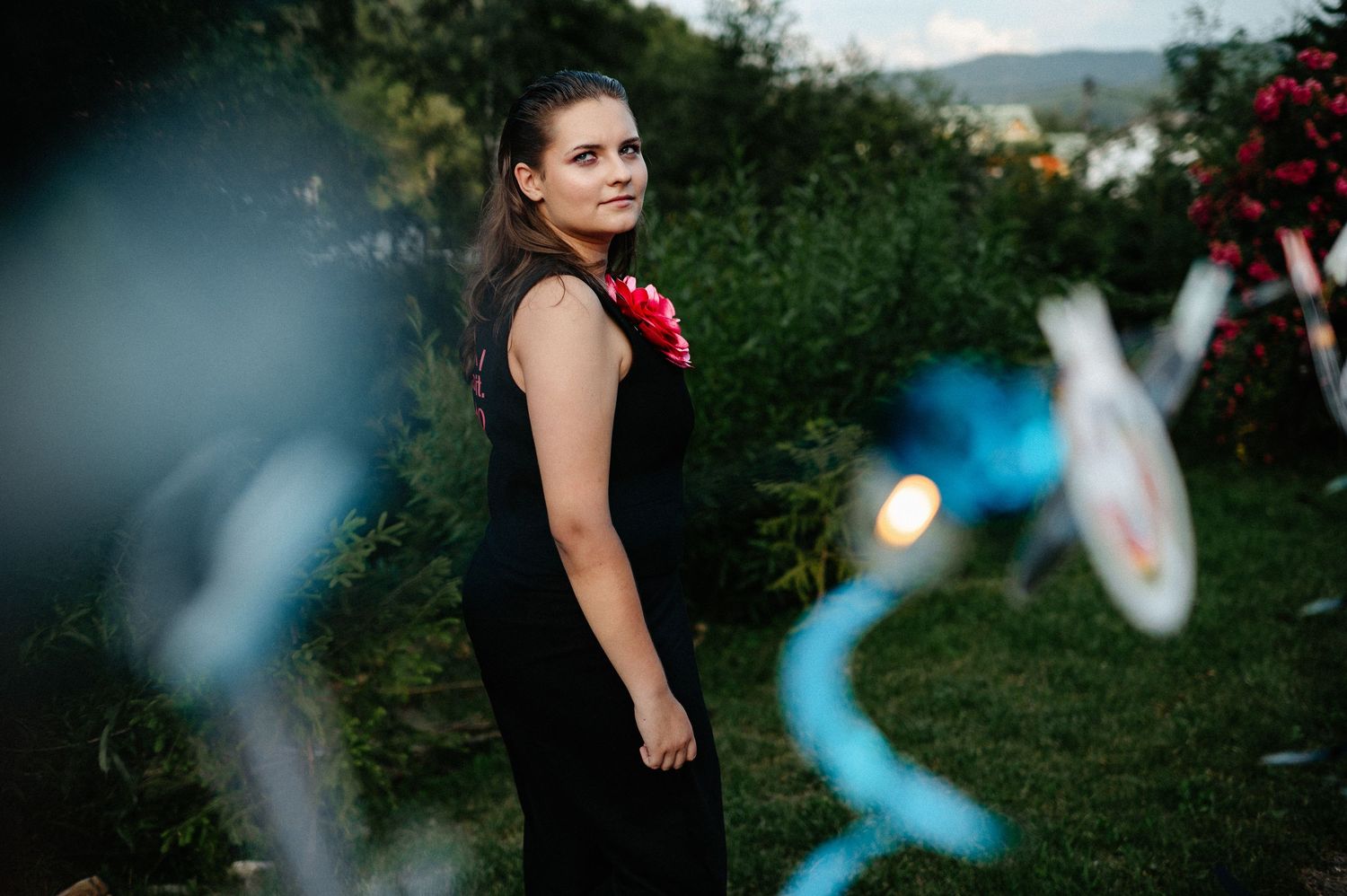 Young woman in black dress with red flower accessory stands in lush garden at dusk.