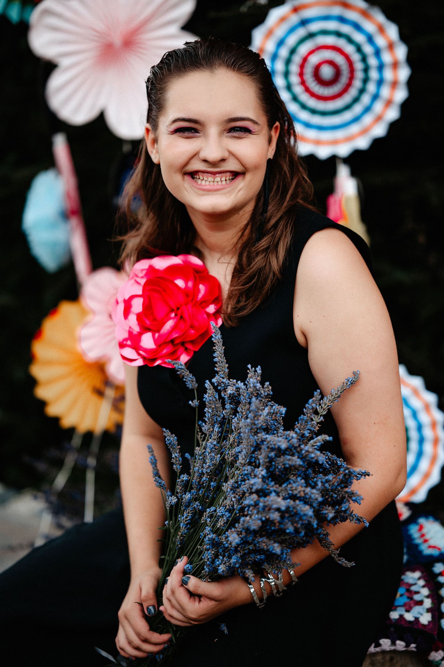 Happy young woman holding lavender bouquet and pink rose corsage with colorful paper fans backdrop.
