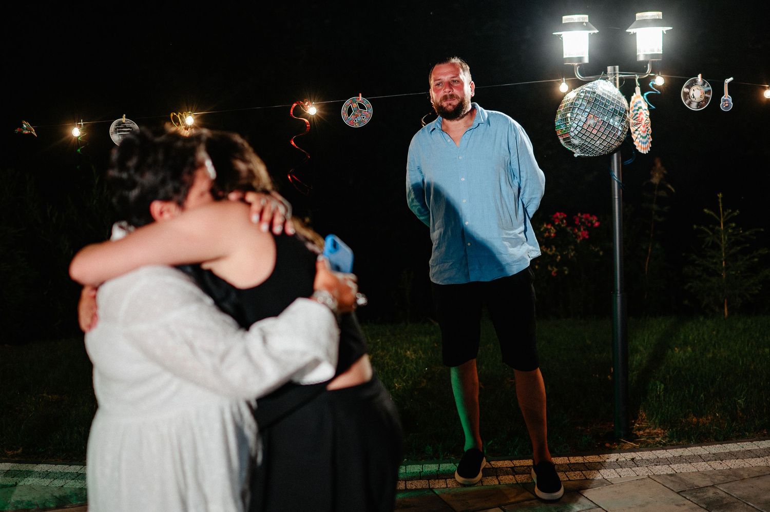 A man watches two women hug at a festive outdoor night party with string lights.