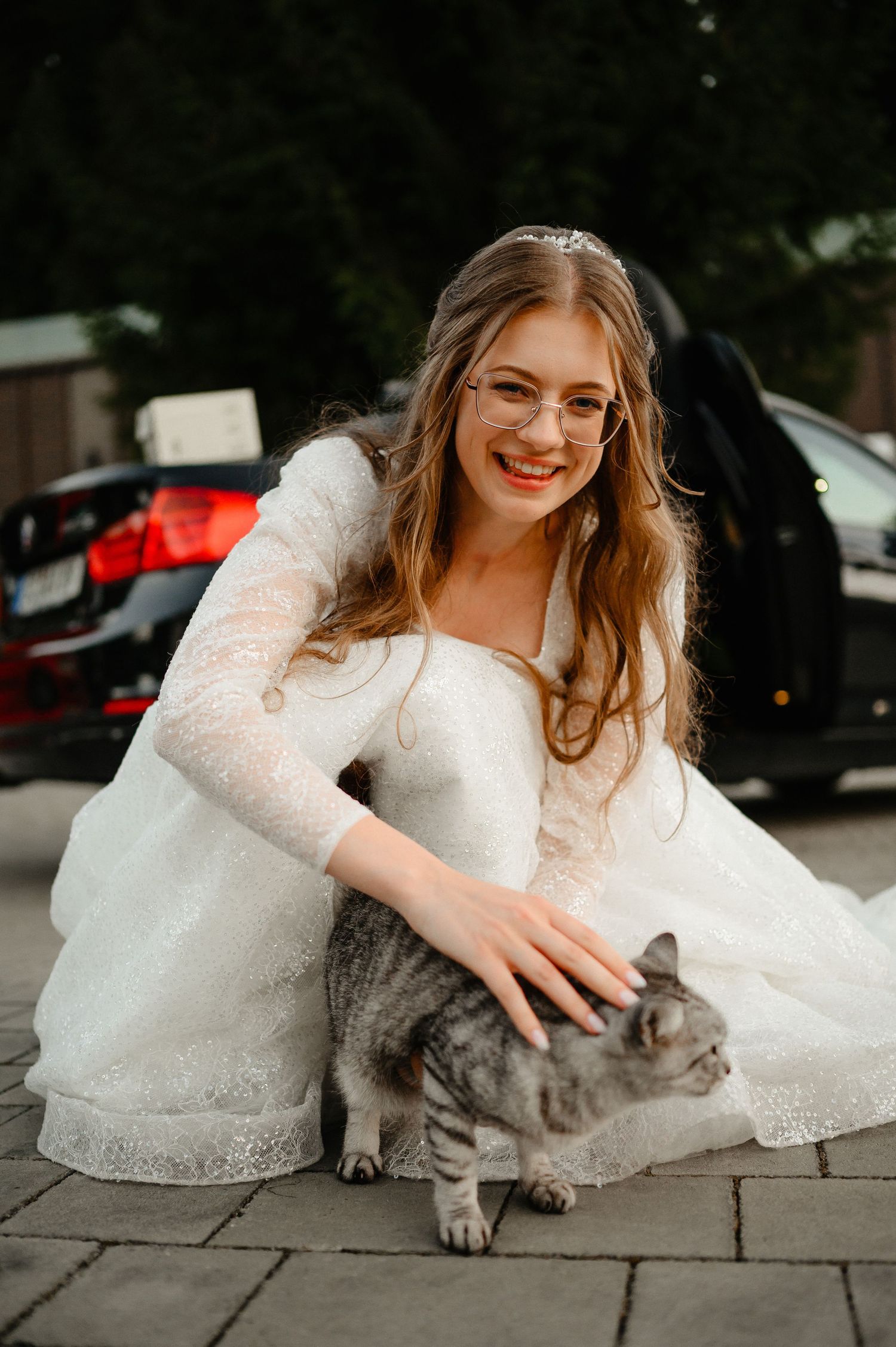 Smiling bride in a lace wedding gown crouches down to pet a tabby cat outdoors.