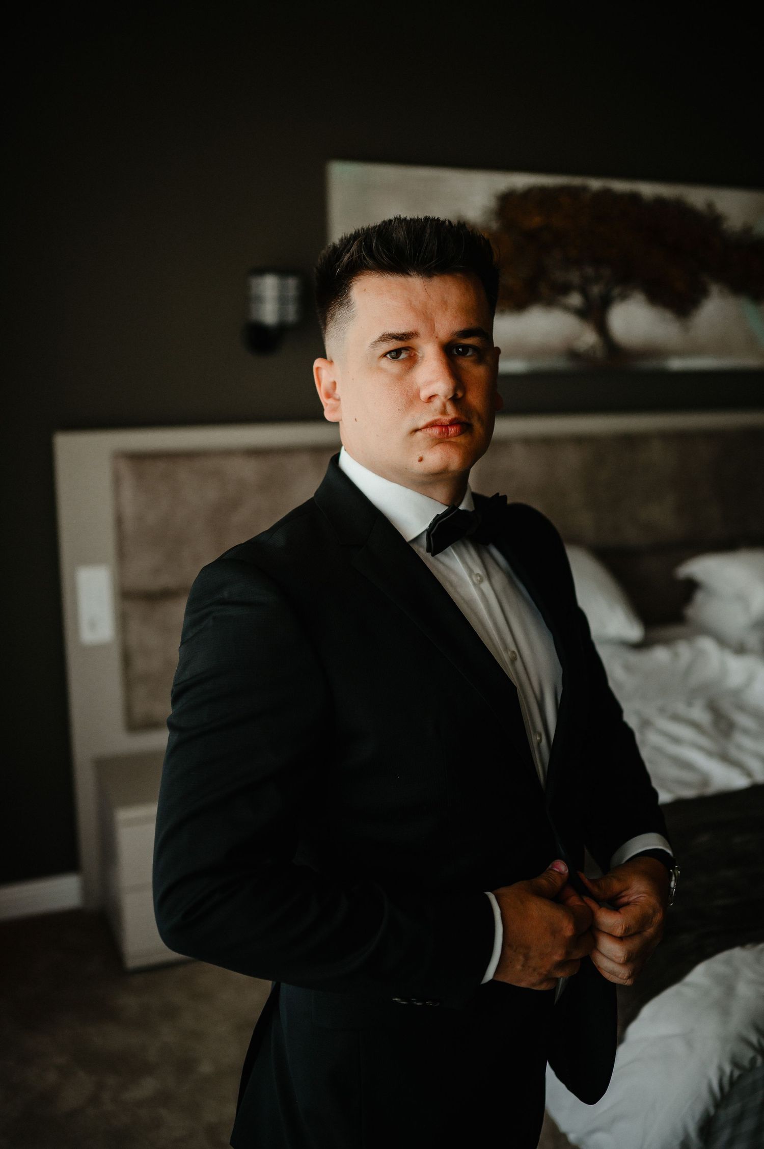 Elegant groom in black tuxedo and bow tie buttoning jacket in stylish dark hotel room.
