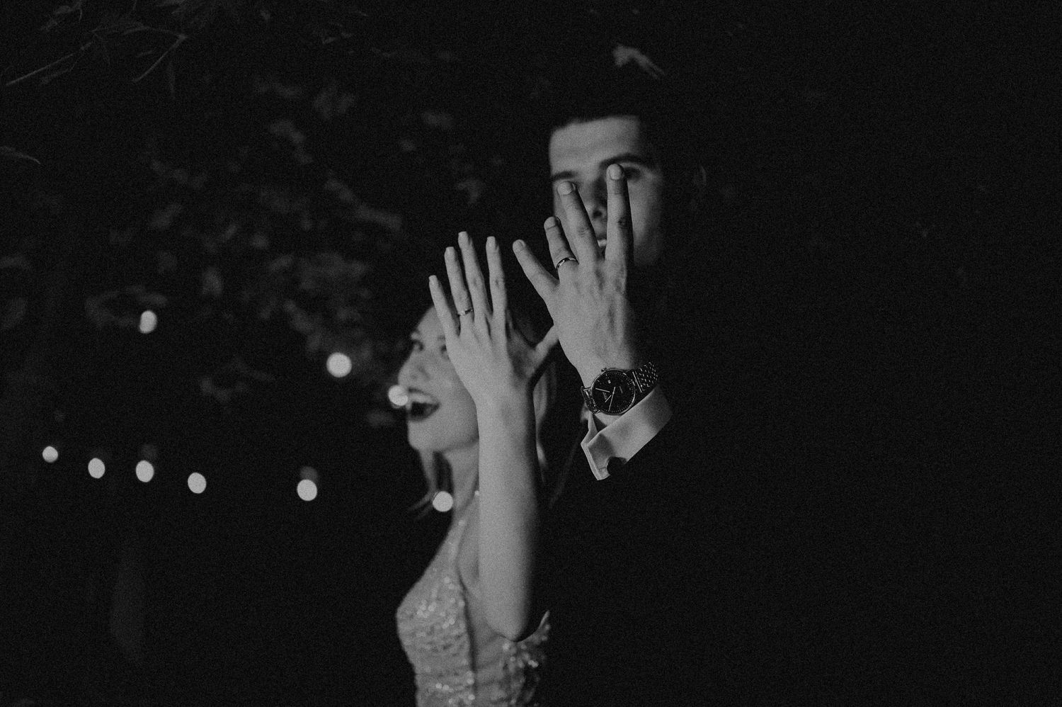 Black and white photo of a couple showing their wedding rings while embracing outdoors at night.