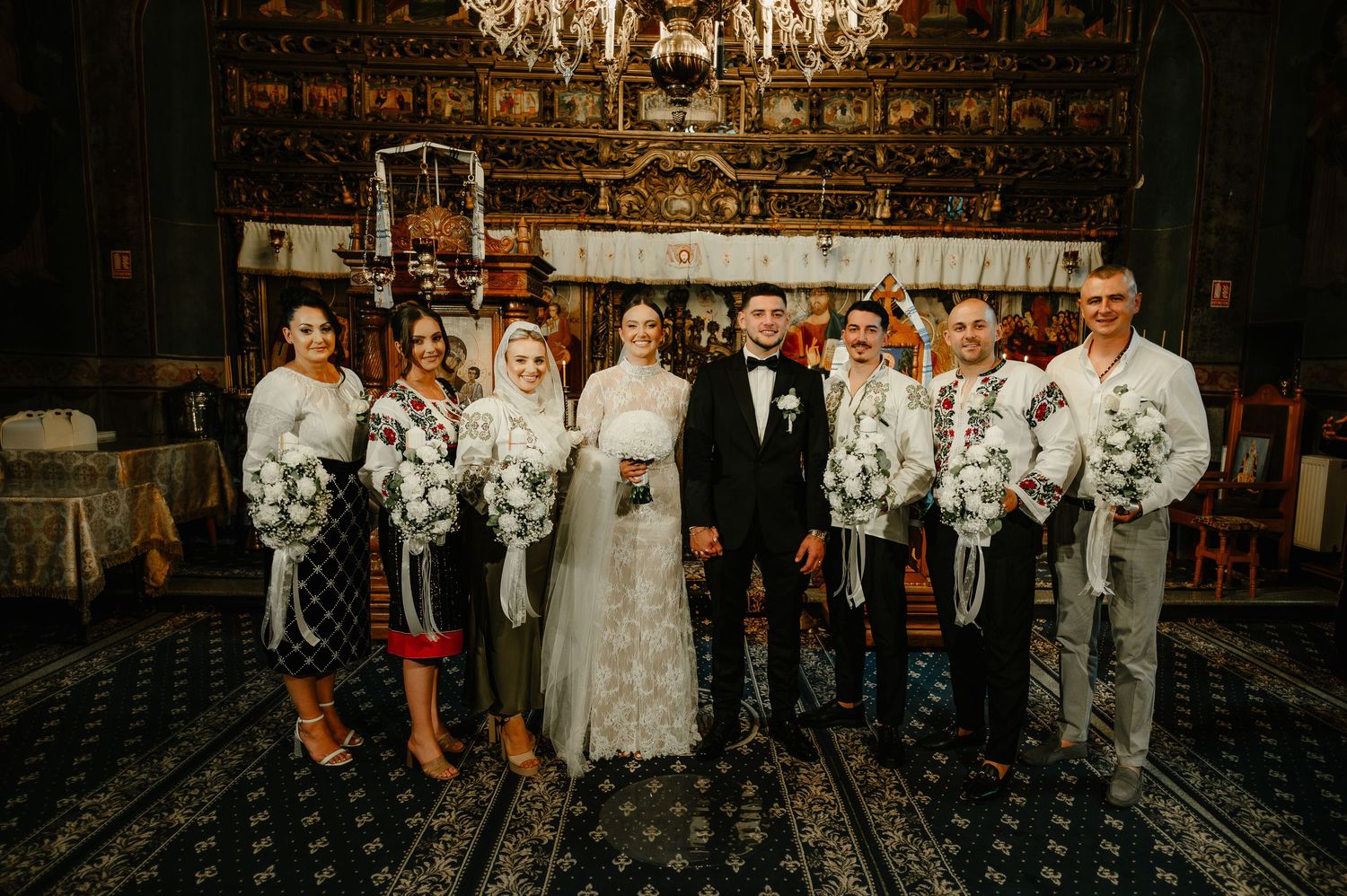 Wedding party in traditional embroidered attire holding white bouquets inside an ornate Orthodox church.