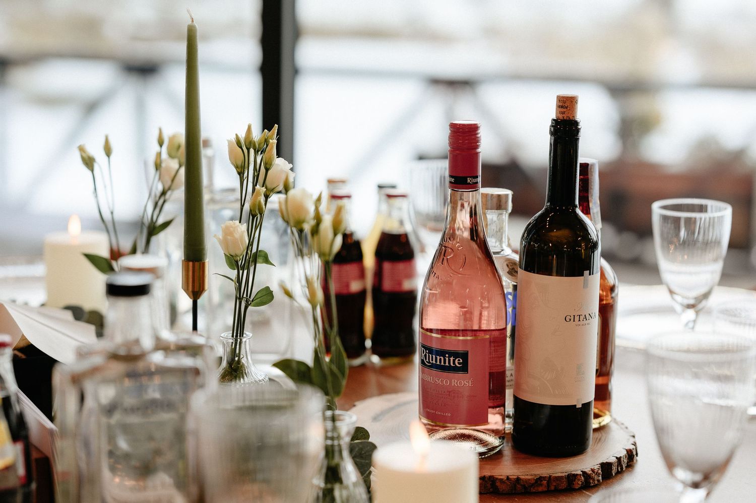 Elegant table setting with wine bottles, pink roses, candles, and glassware on a wooden tray.
