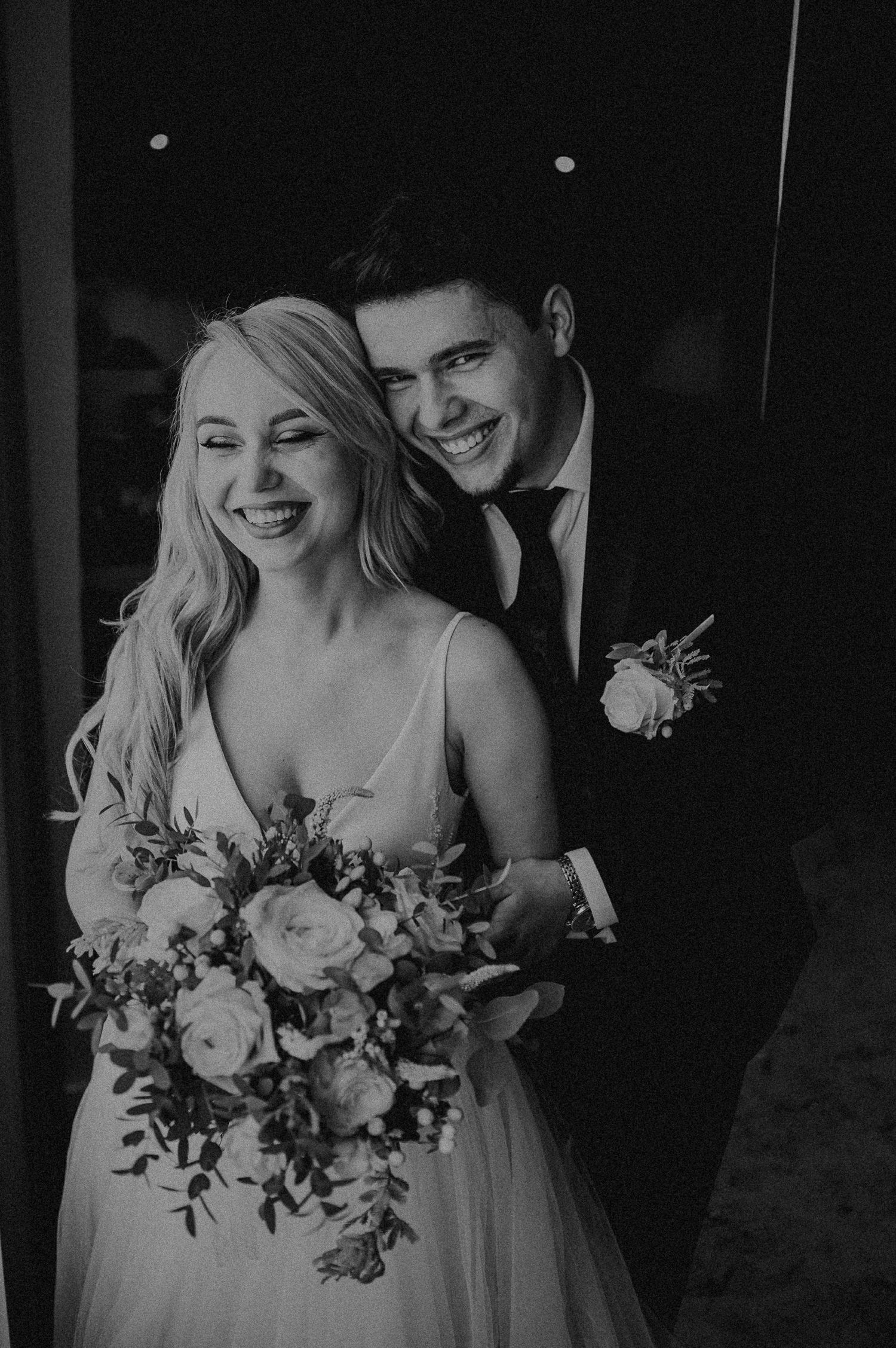 Black and white portrait of laughing bride and groom embracing with white floral bouquet.