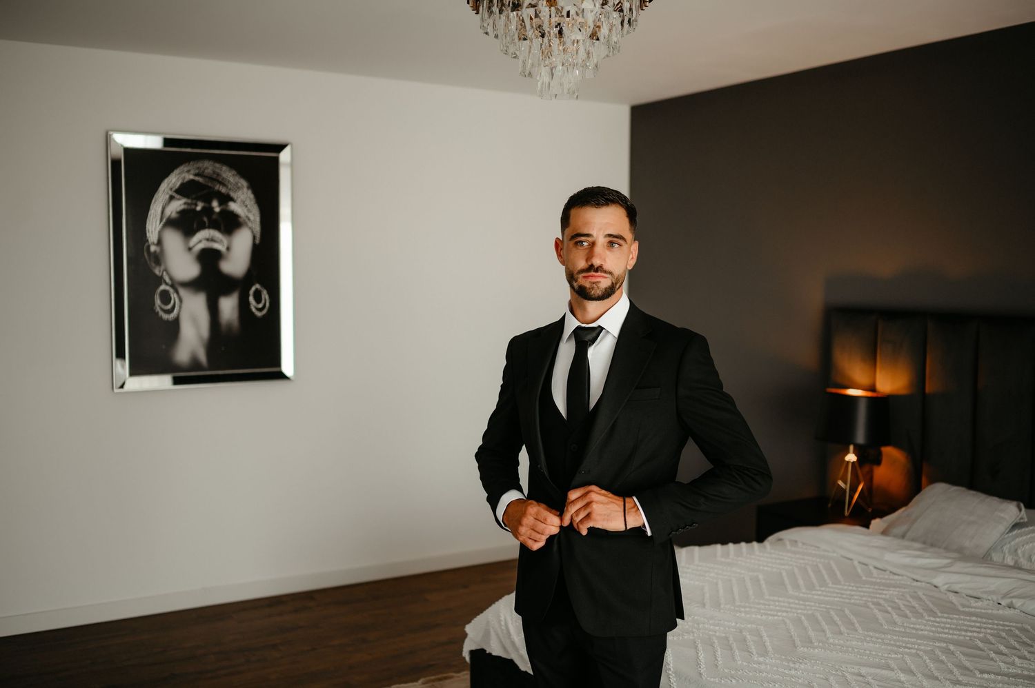 Groom in black suit buttoning jacket in elegant hotel bedroom.