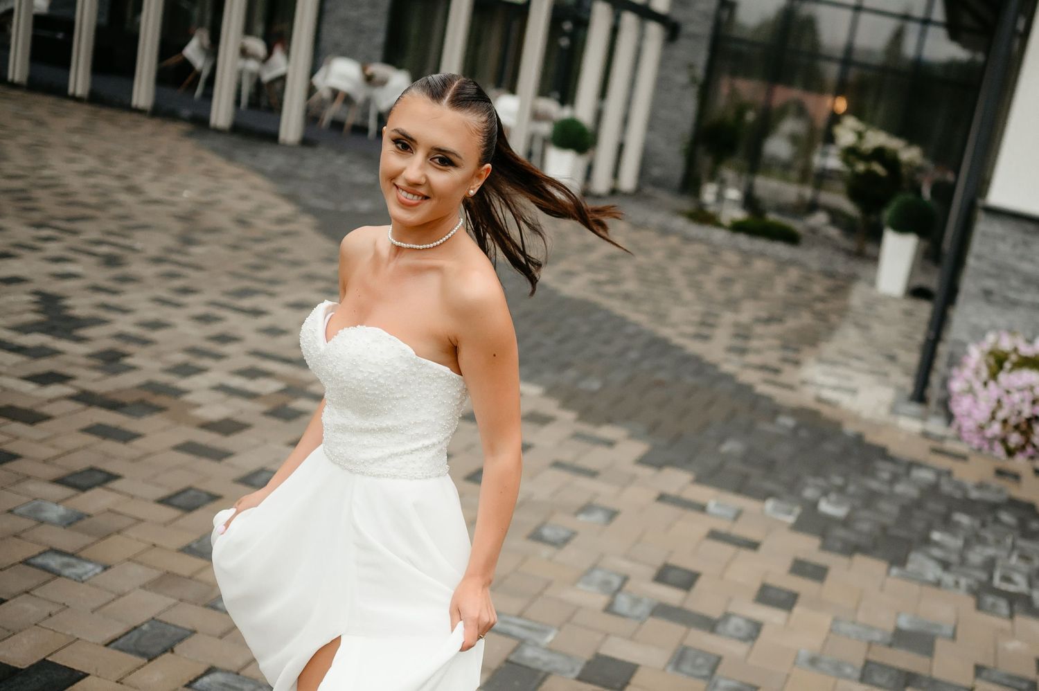 Smiling bride in strapless white dress twirling on a cobblestone courtyard with flowers in background.