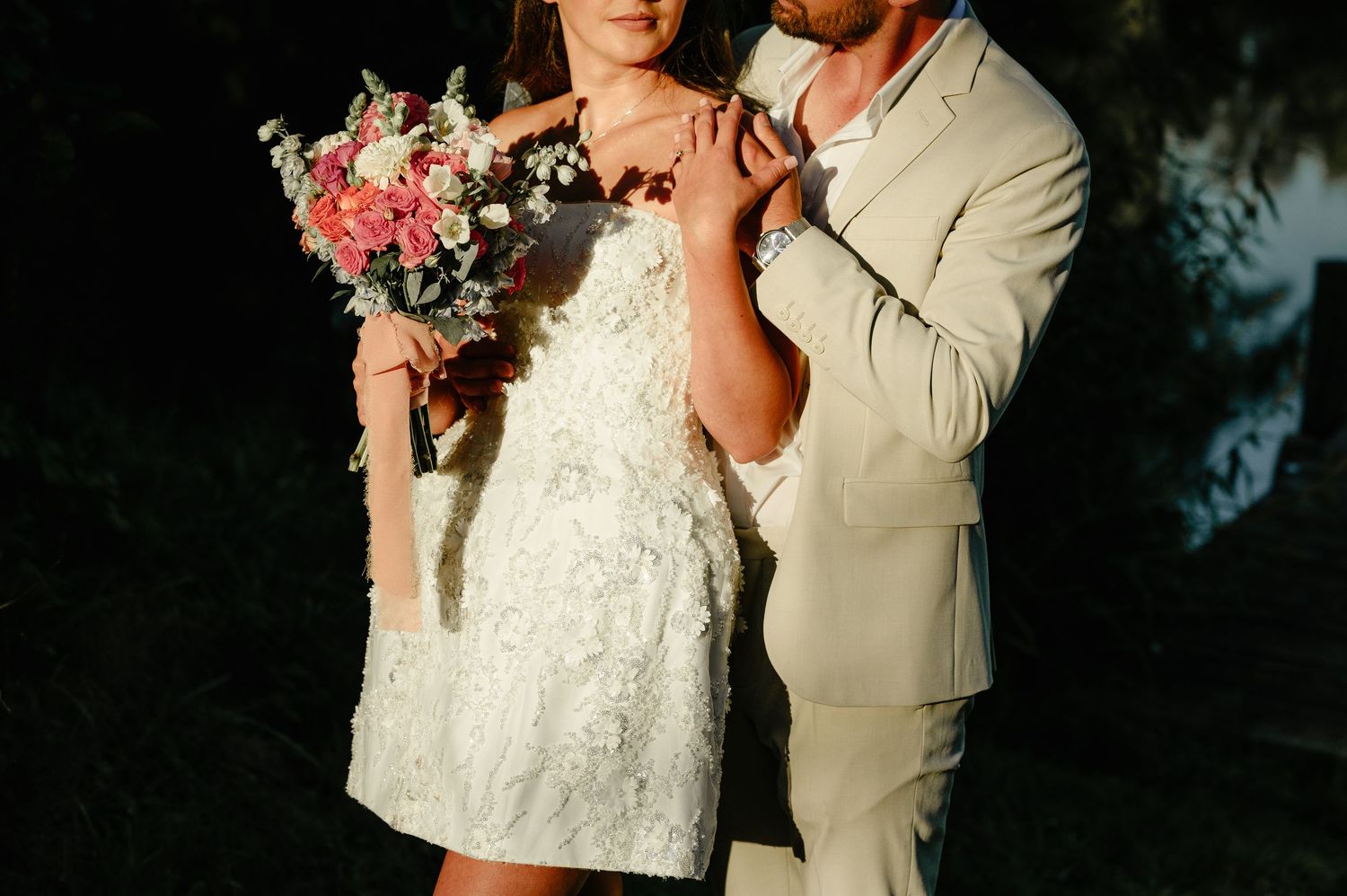 Bride and groom embracing at sunset, bride in short white lace dress holding pink floral bouquet.