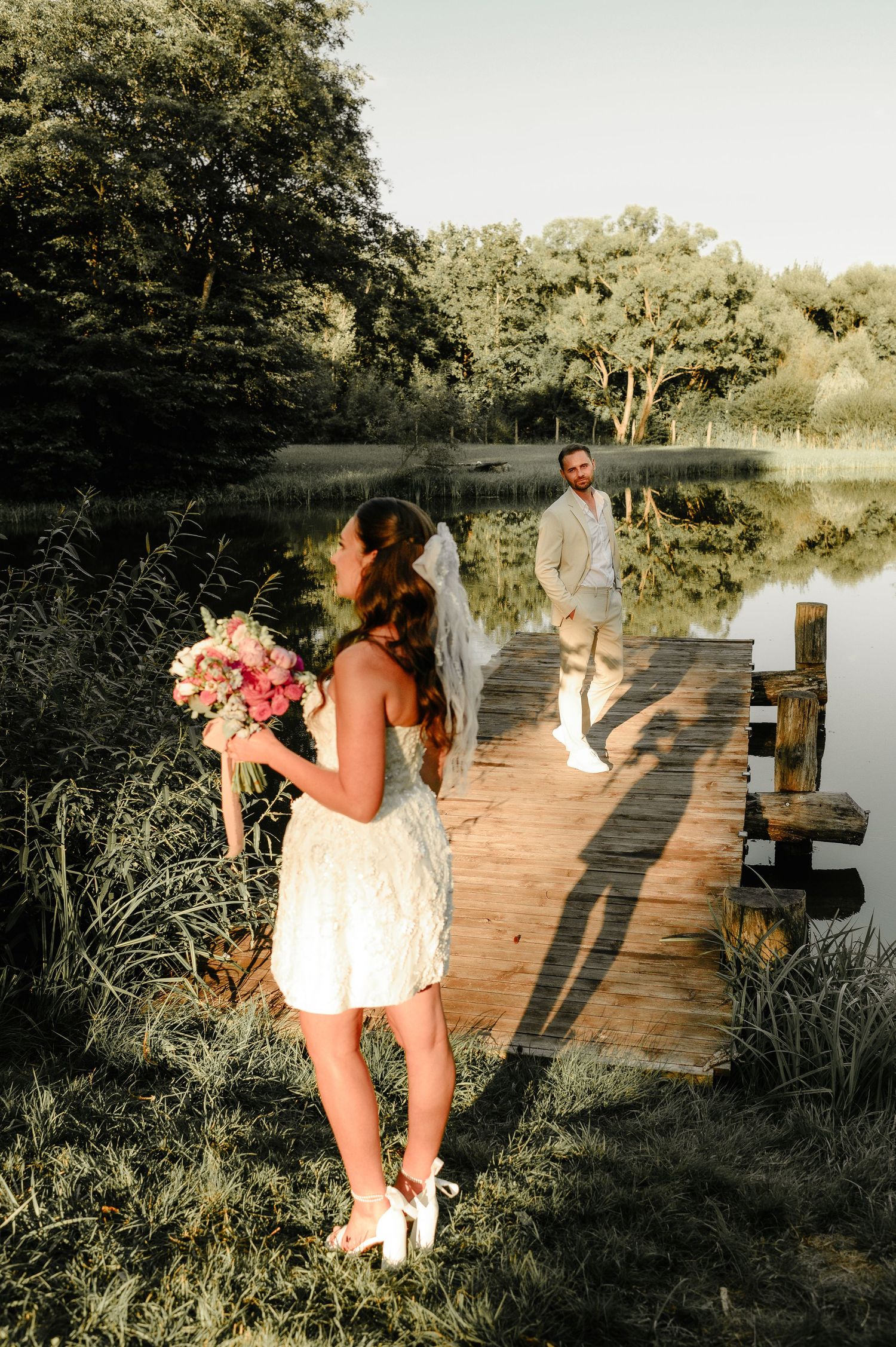 Bride in short white lace dress holds pink bouquet on wooden dock beside a serene reflective lake.