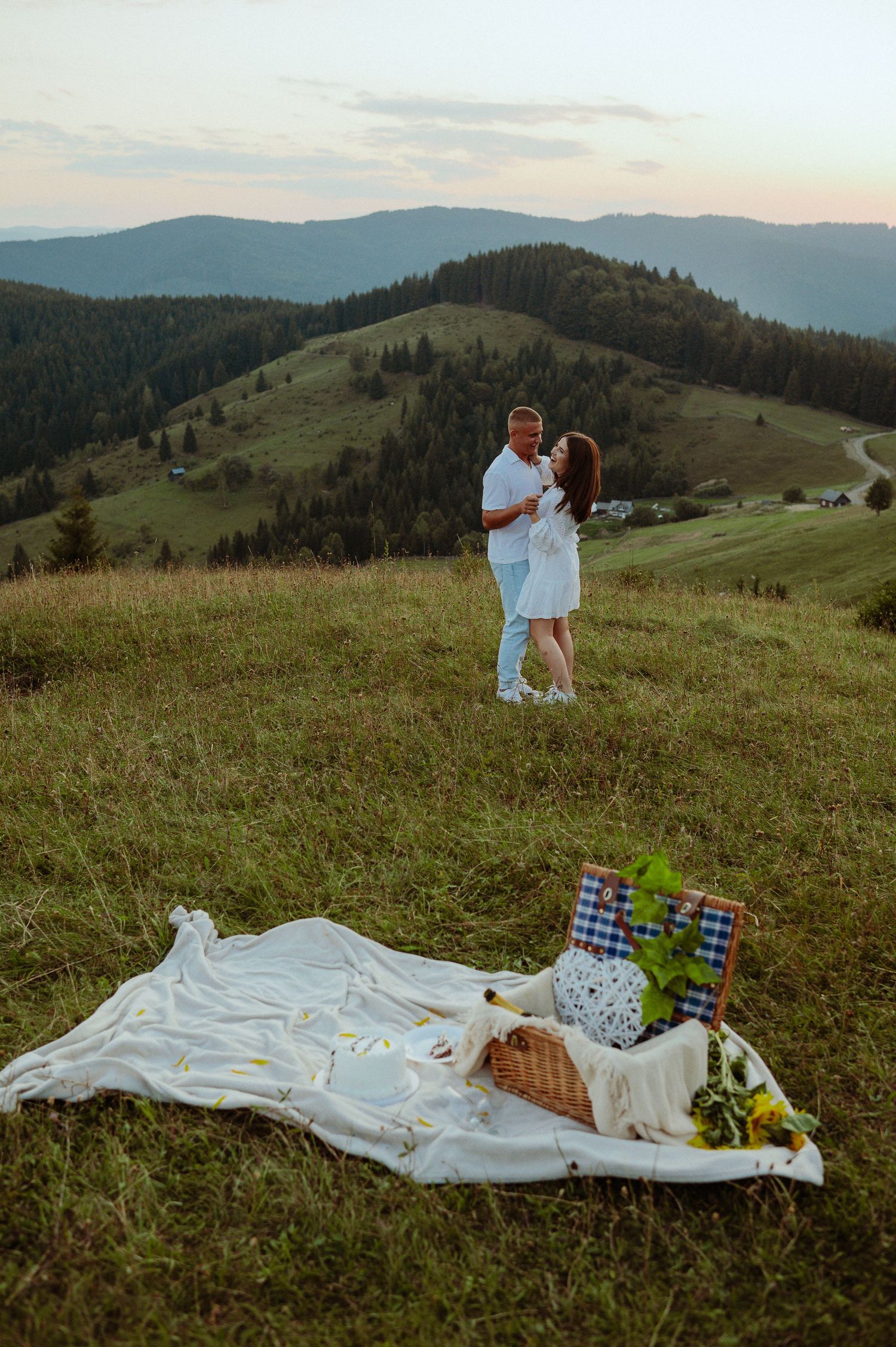 Romantic couple dancing on mountain hillside at sunset near picnic blanket with basket and flowers.