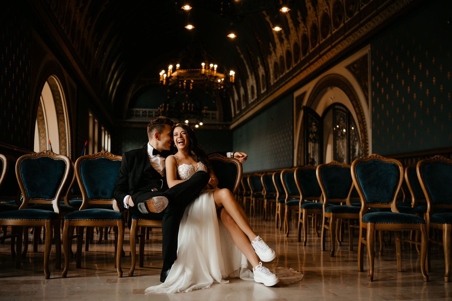 Groom holds bride in his arms while sitting together in an elegant ornate ballroom with chandeliers.