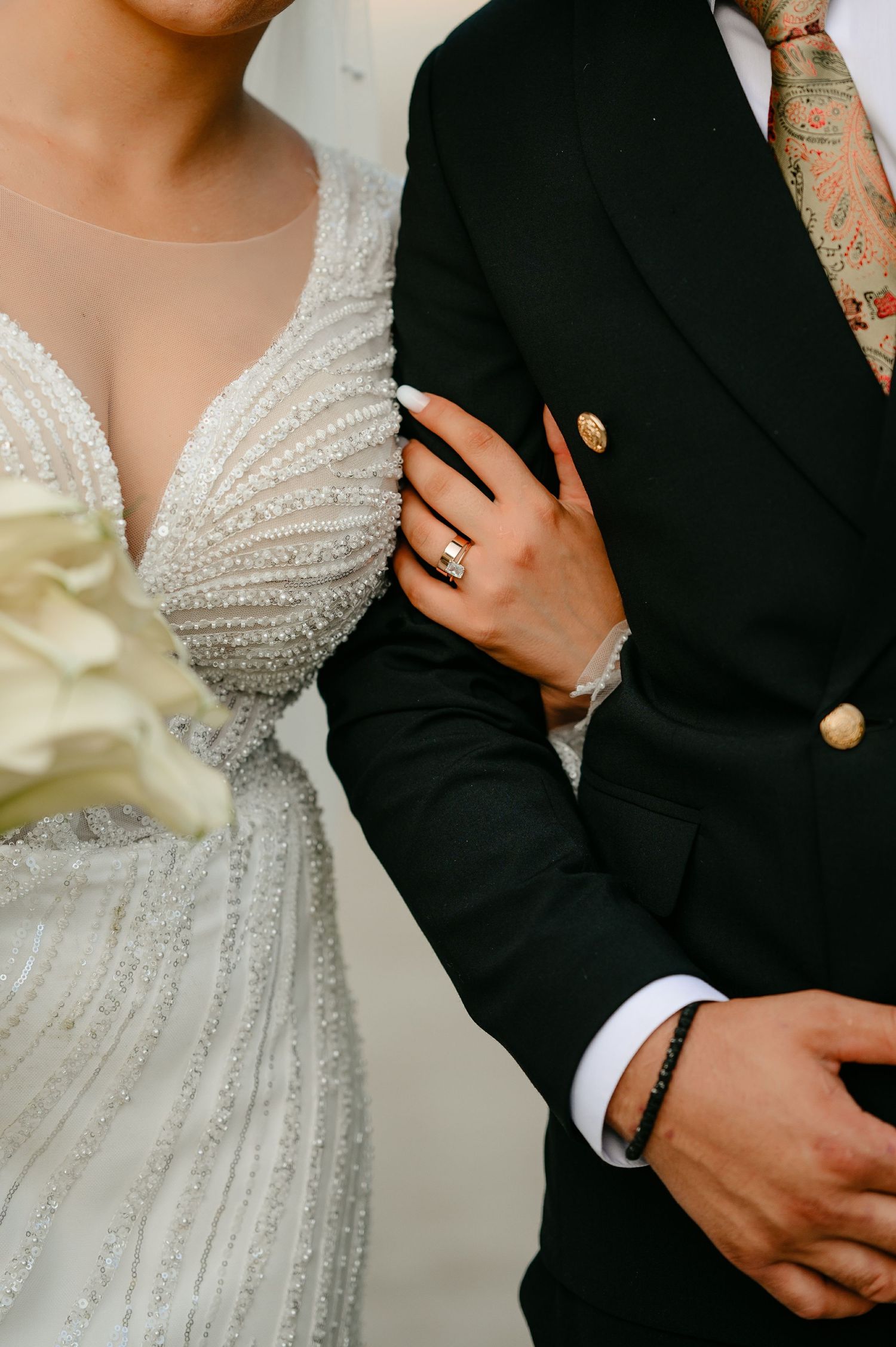 Close-up of bride in beaded wedding dress holding groom's arm, showcasing diamond engagement ring and bouquet.