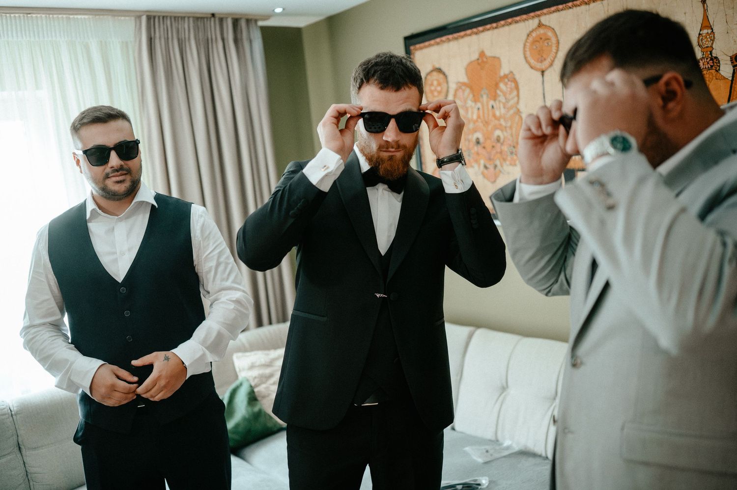 Three men in formal suits trying on sunglasses together while getting ready for a wedding.
