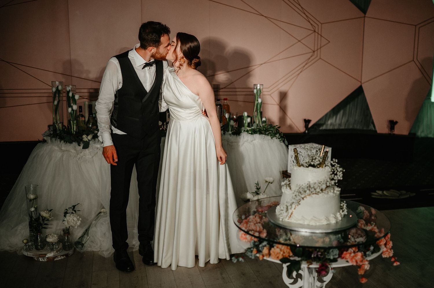 Newlyweds share a tender kiss beside their white wedding cake at their elegant reception venue.
