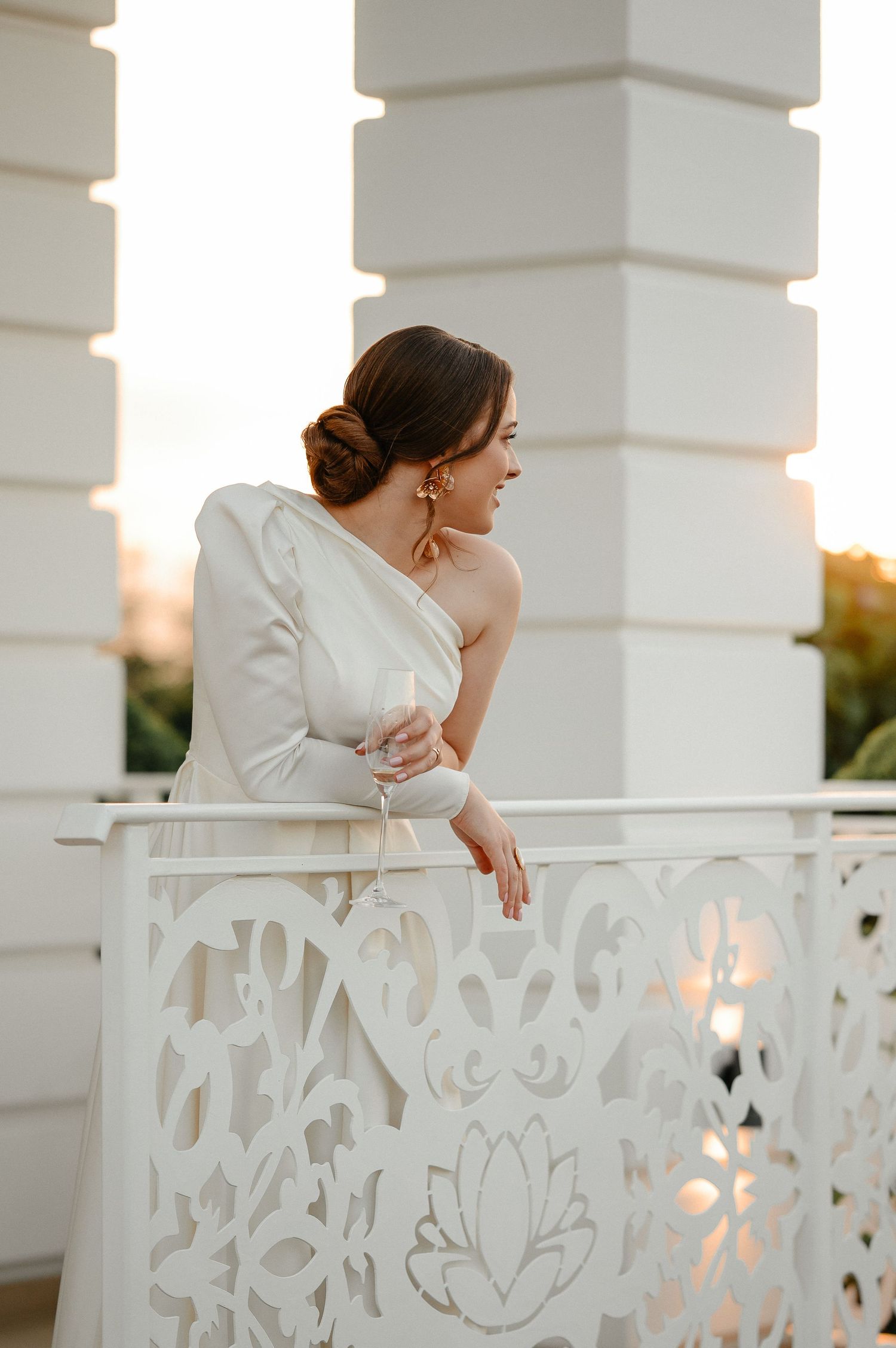 Elegant bride in white one-shoulder gown stands on decorative white balcony at golden sunset.