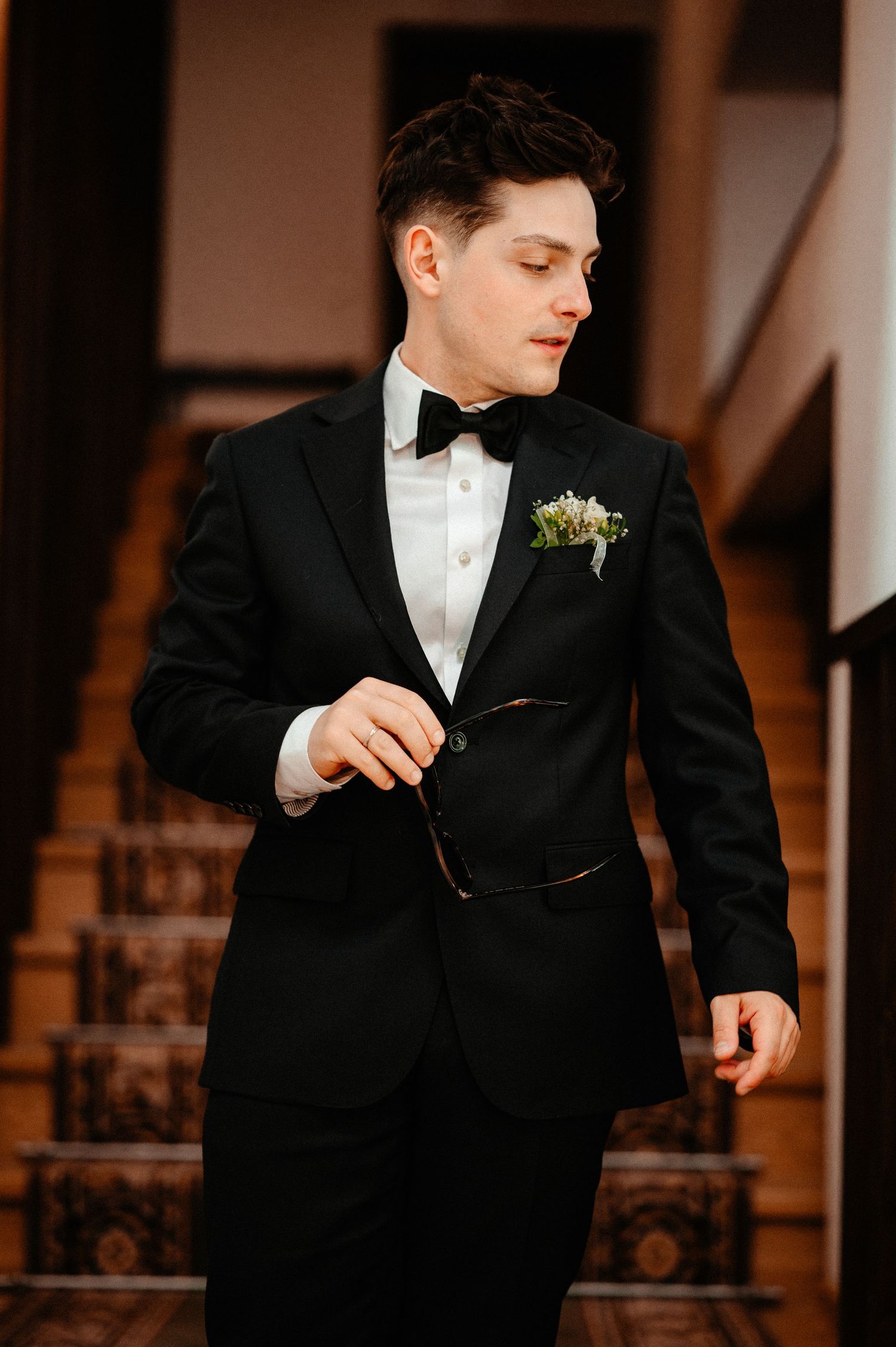 Groom in black tuxedo with bow tie and boutonniere holding glasses near staircase.