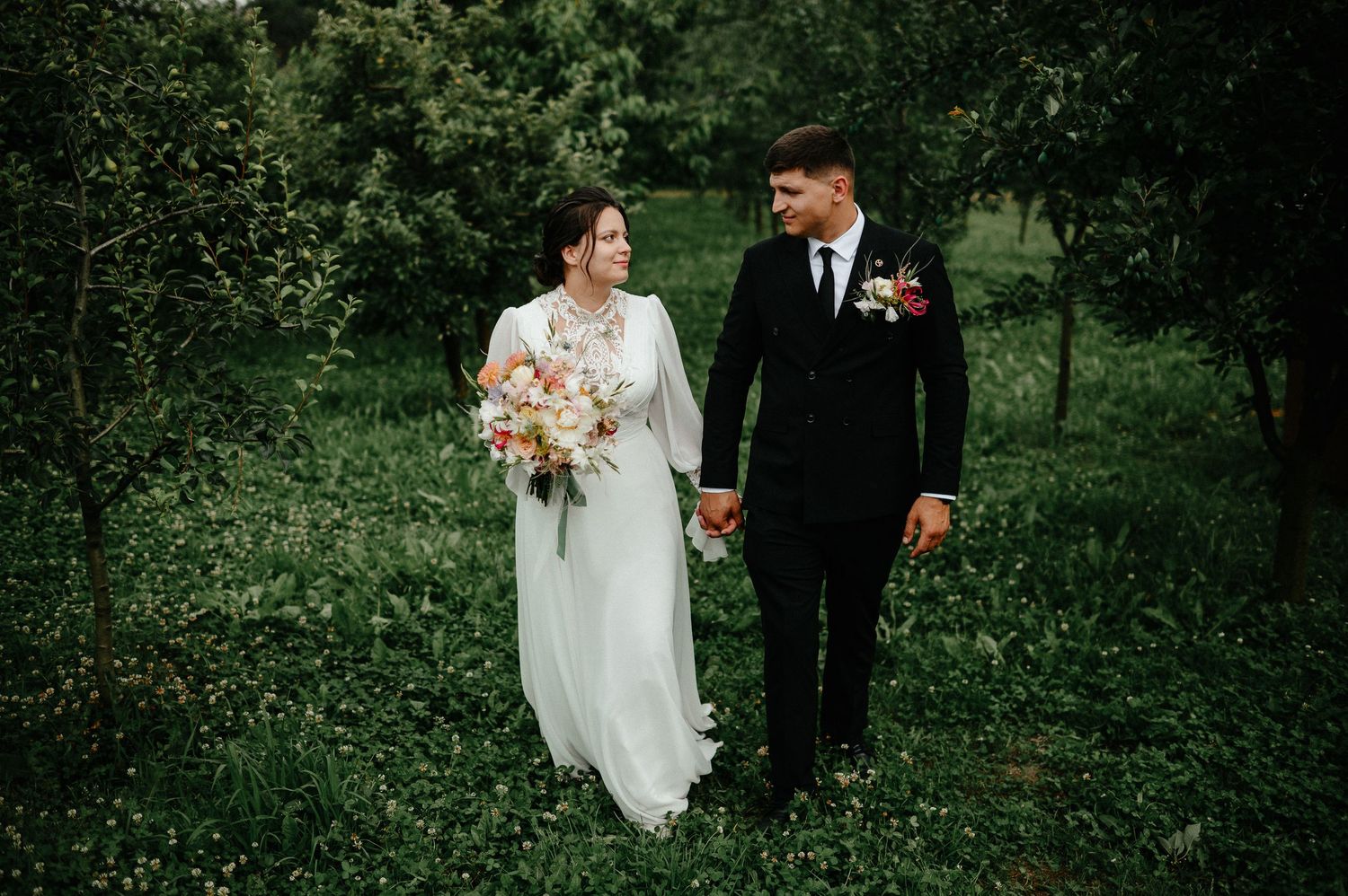 Bride and groom walking hand in hand through lush green garden with floral bouquet.