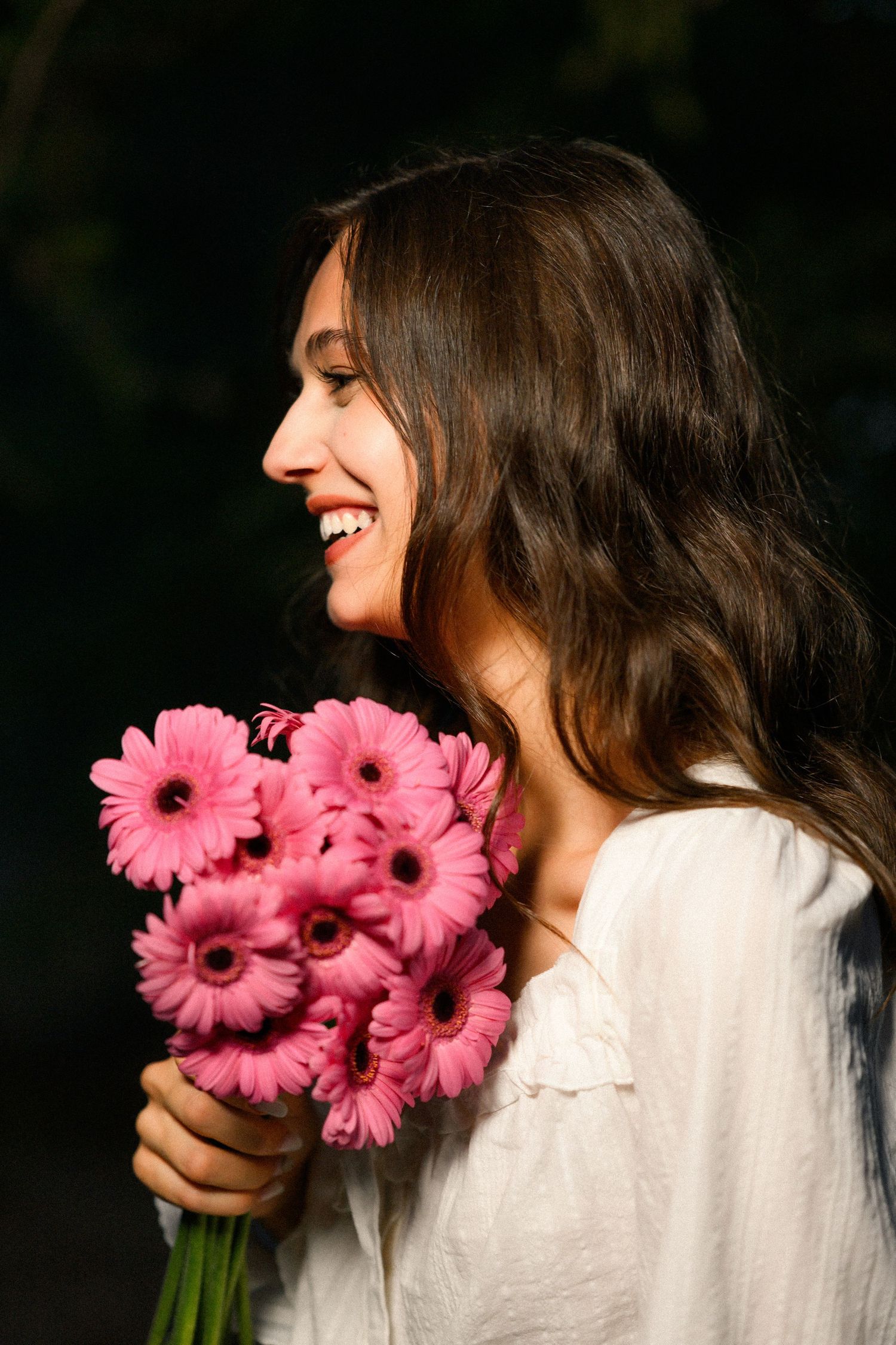 Smiling young woman holding a bouquet of pink gerbera daisies, wearing a white blouse outdoors.