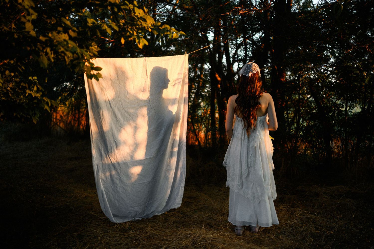 Woman in white dress stands before a sheer fabric with a shadowy silhouette in a mystical forest at dusk.