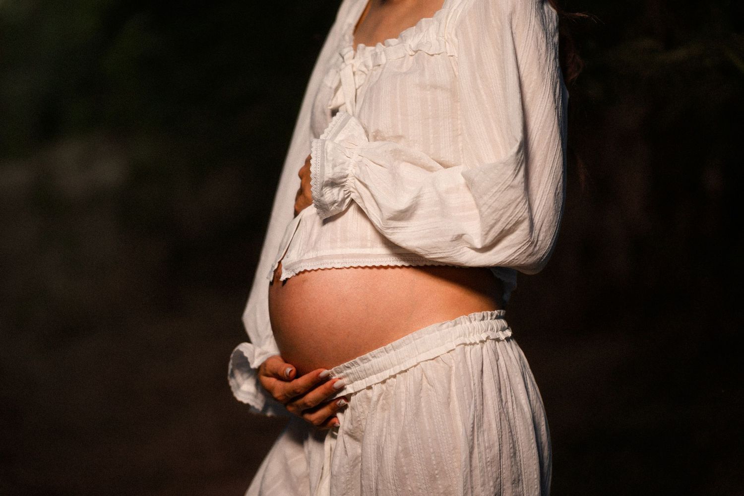 Pregnant woman in white lace dress cradling her bare baby bump outdoors in soft natural light.