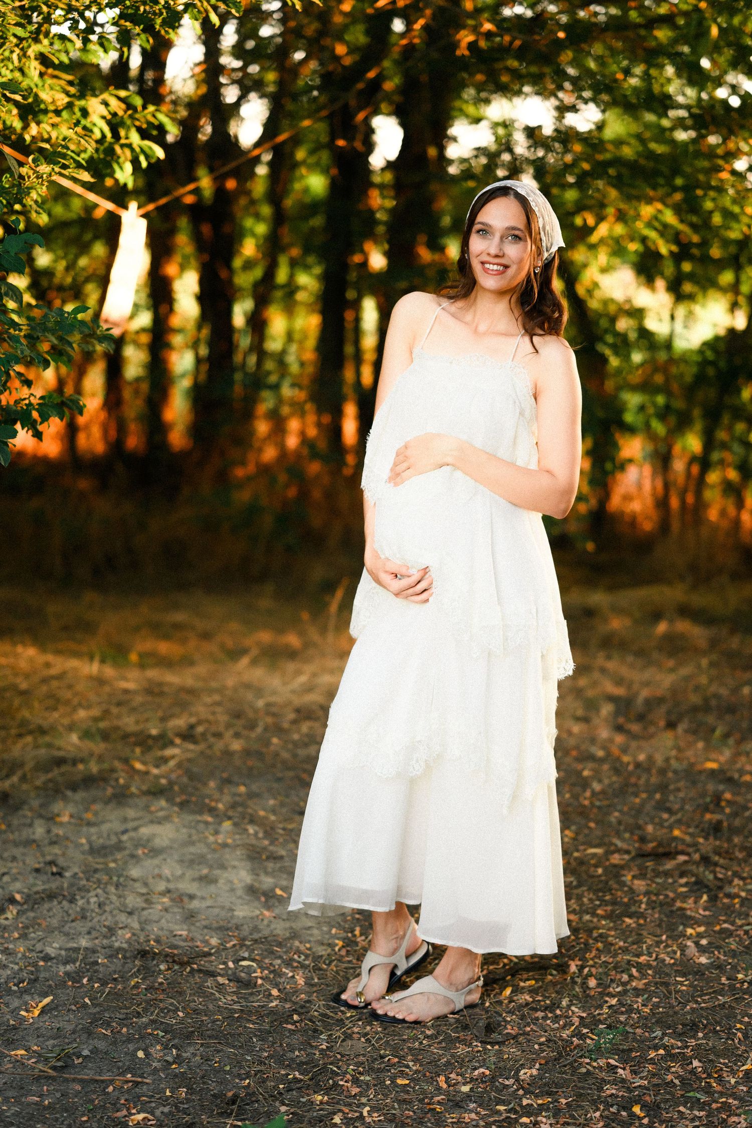 Smiling pregnant woman in white sundress standing outdoors in golden sunlight among autumn trees.