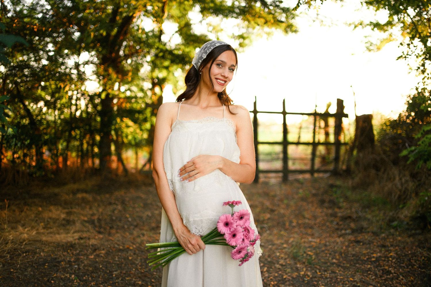 Smiling pregnant woman in white dress holding pink flowers outdoors in golden sunlight near rustic fence.
