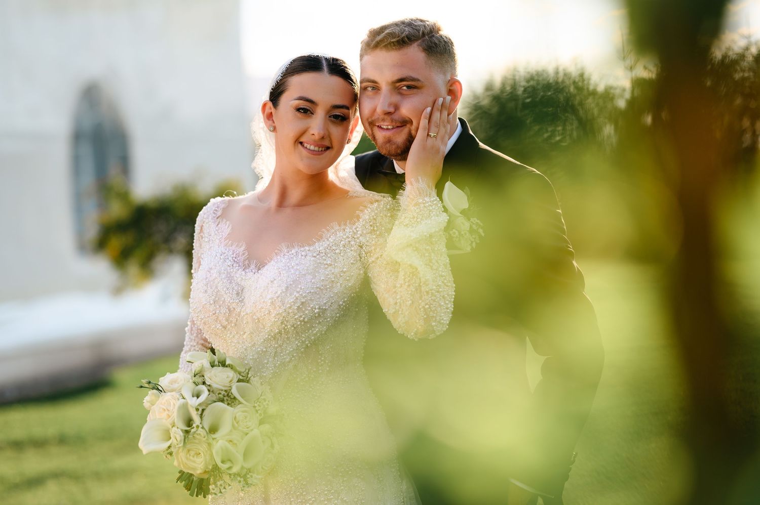 Bride in lace gown holding white bouquet poses with groom in suit at outdoor wedding ceremony.