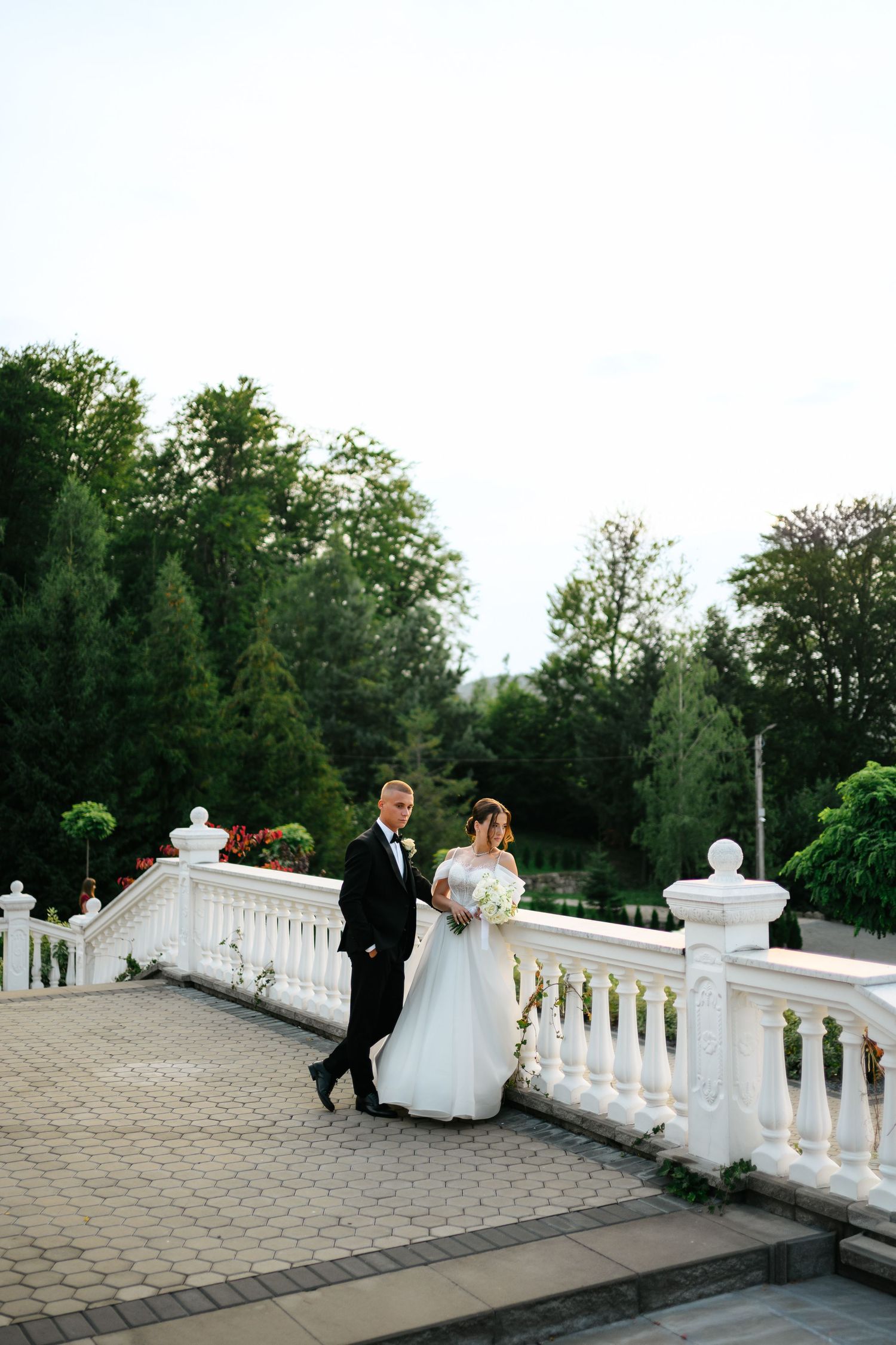 Bride and groom walking on elegant white balustrade terrace surrounded by lush green trees on their wedding day.