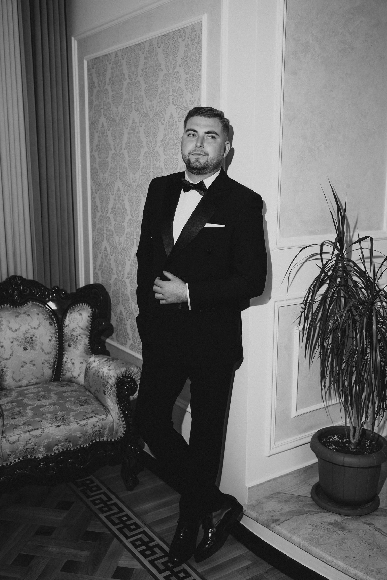 Black and white photo of a well-dressed man in a tuxedo posing elegantly in a classic interior room.