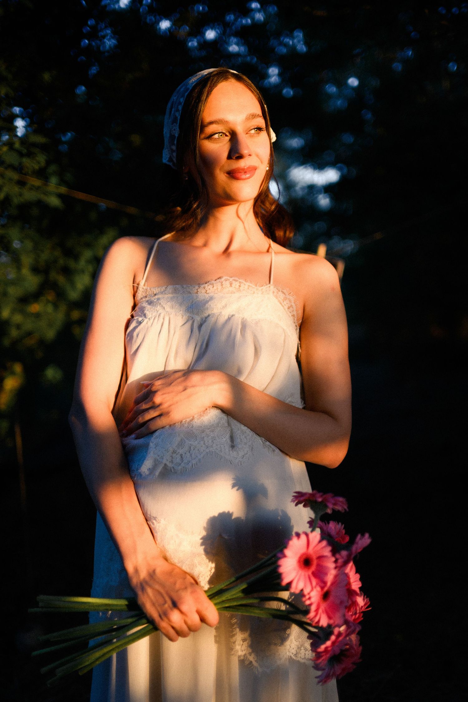 Woman in white lace dress holding pink gerbera daisies, bathed in golden sunlight against a dark garden background.