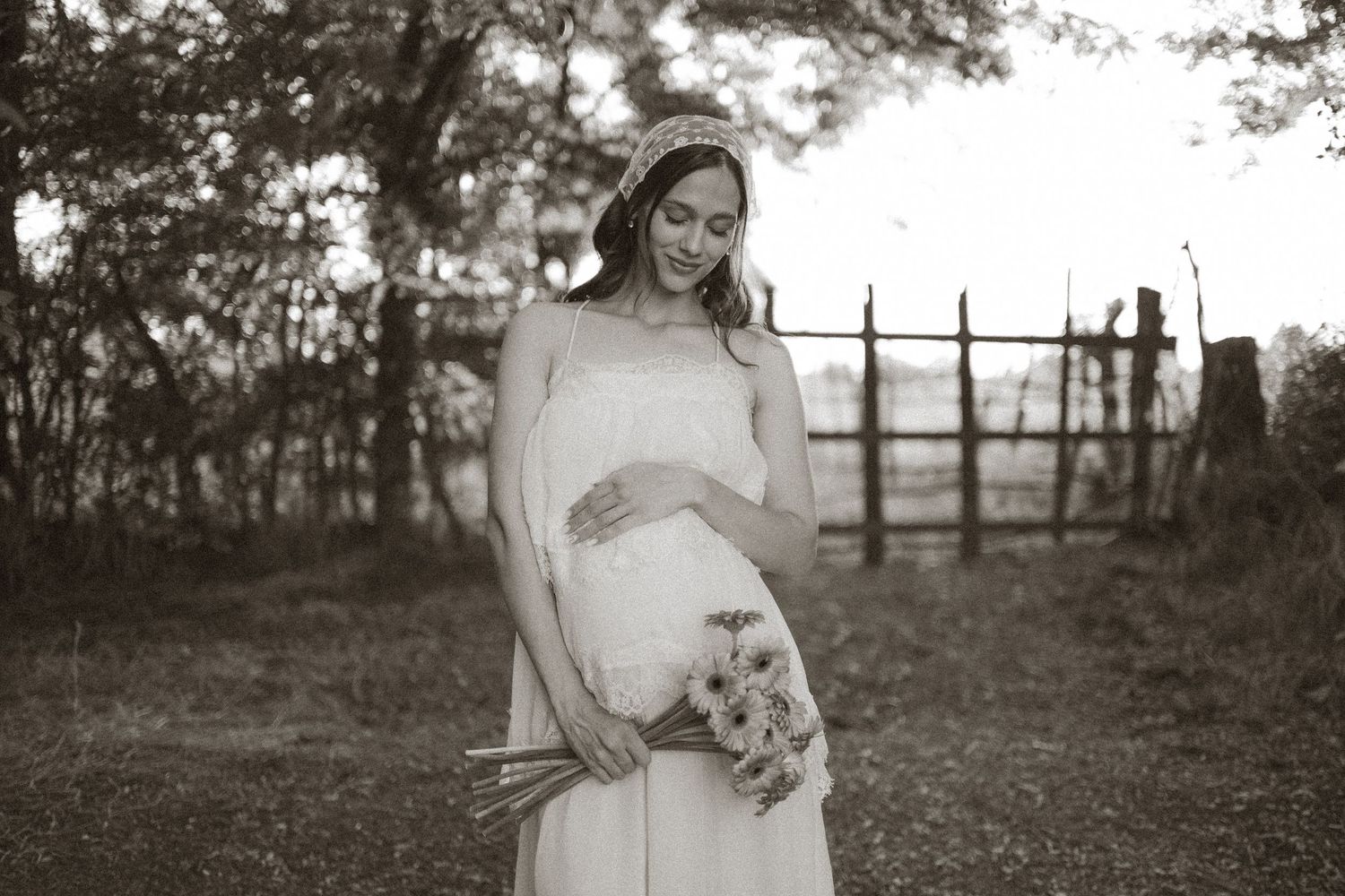 Pregnant bride in white gown holding bouquet, smiling outdoors in black and white maternity photo.