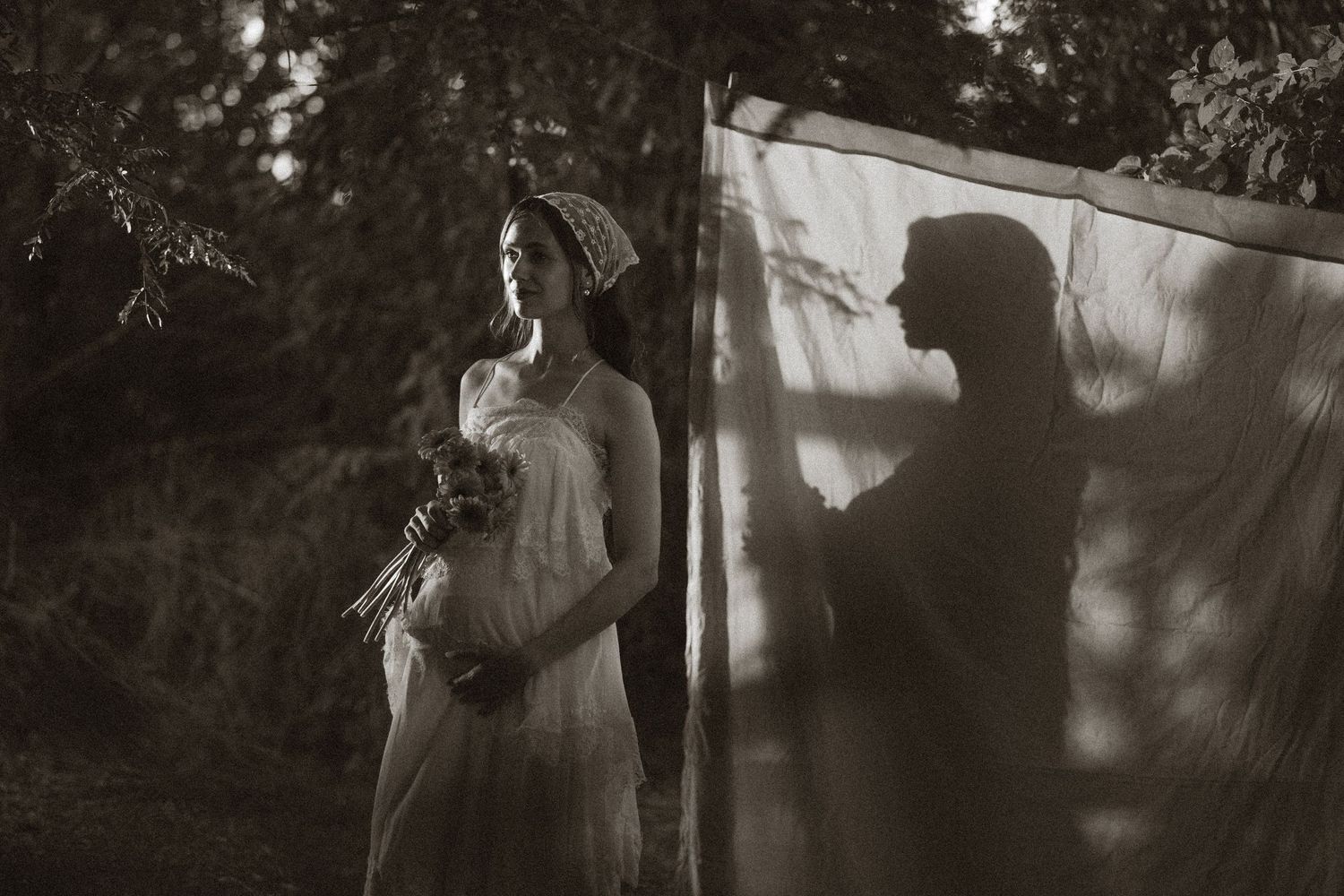 Pregnant woman in white dress holding flowers outdoors, with a silhouette shadow cast on a sheer fabric backdrop.