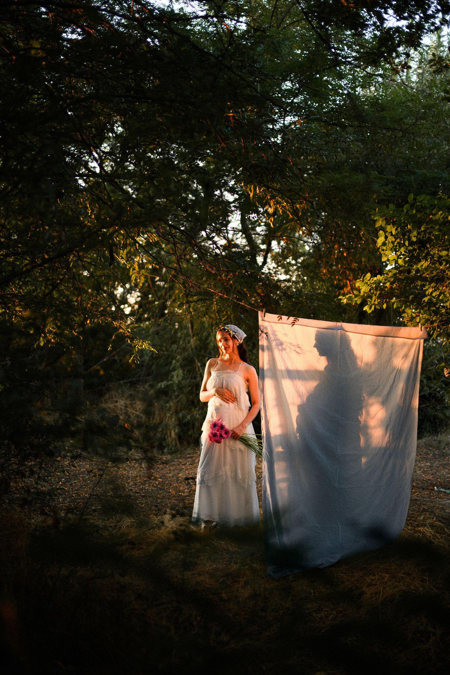 Bride in white gown holding pink bouquet stands in sunlit forest beside flowing fabric with shadow silhouette.