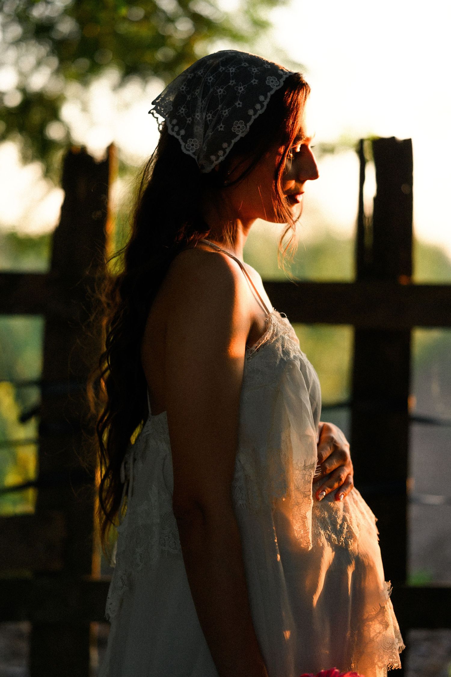 Pregnant woman in white dress and lace headscarf stands in golden sunset light, cradling her baby bump outdoors.