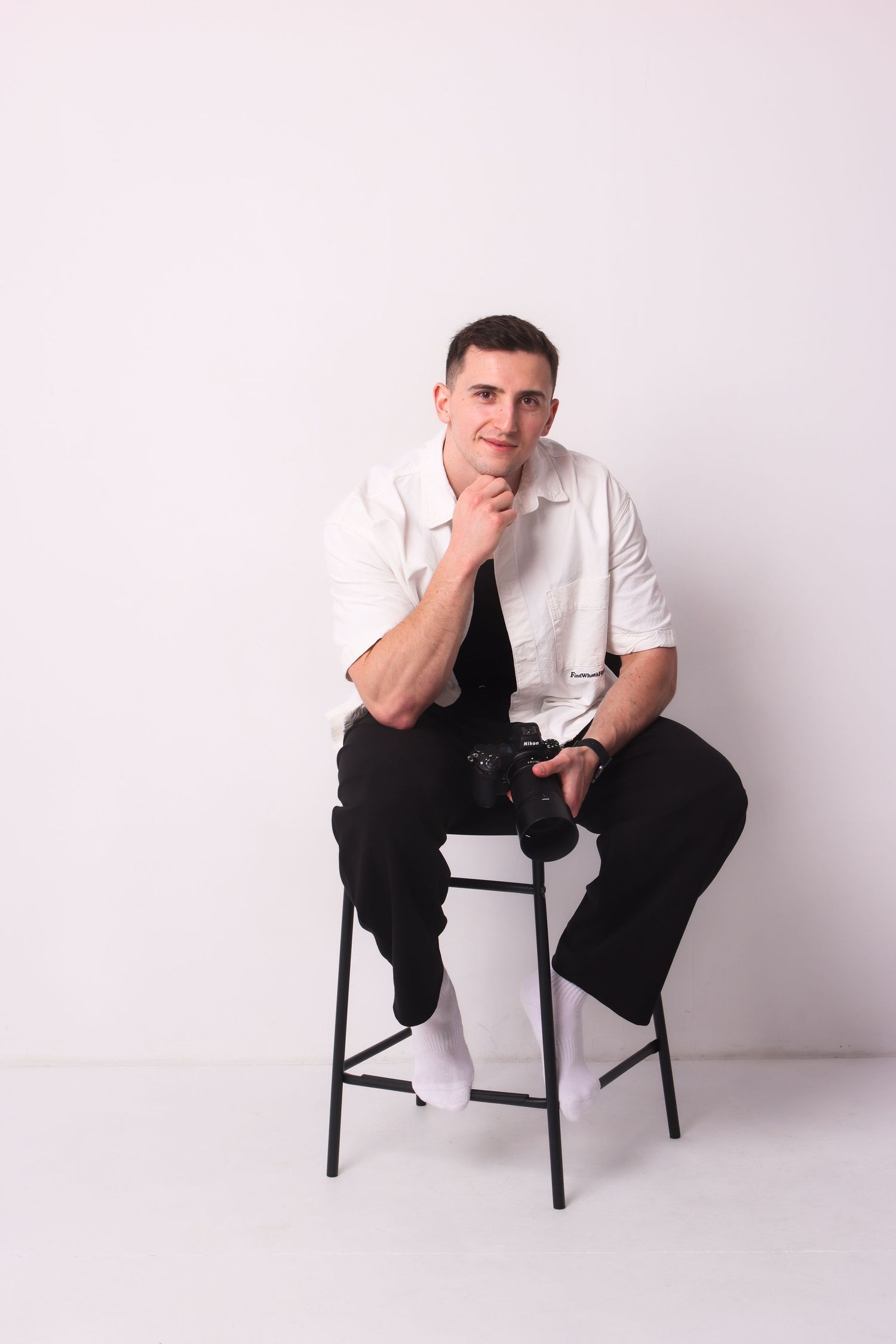 A thoughtful man in white shirt sits on stool holding camera with hand on chin, white background.