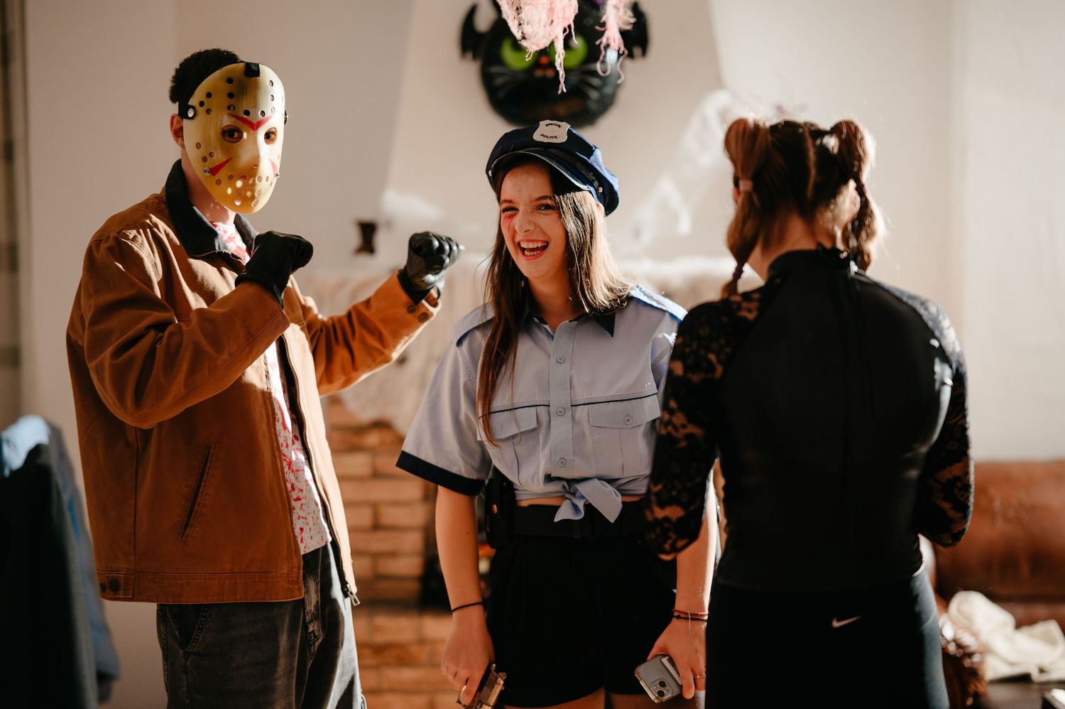 Three people in Halloween costumes including Jason mask and police uniform laugh at indoor party.